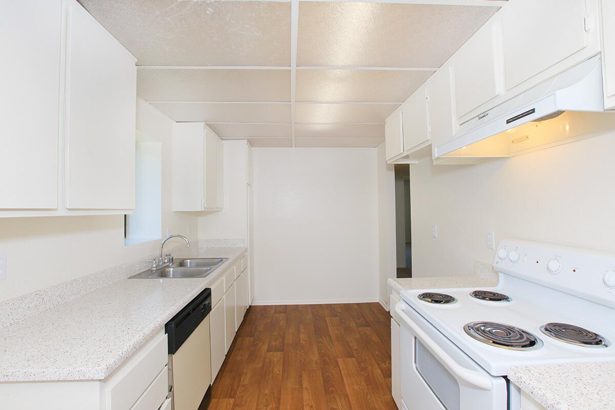Interior view of a modern kitchen featuring white cabinets, a stainless steel sink, and a white stove with an oven. The kitchen has a light-colored countertop and wooden flooring, with a simple layout and neutral wall colors, creating a clean and spacious atmosphere.