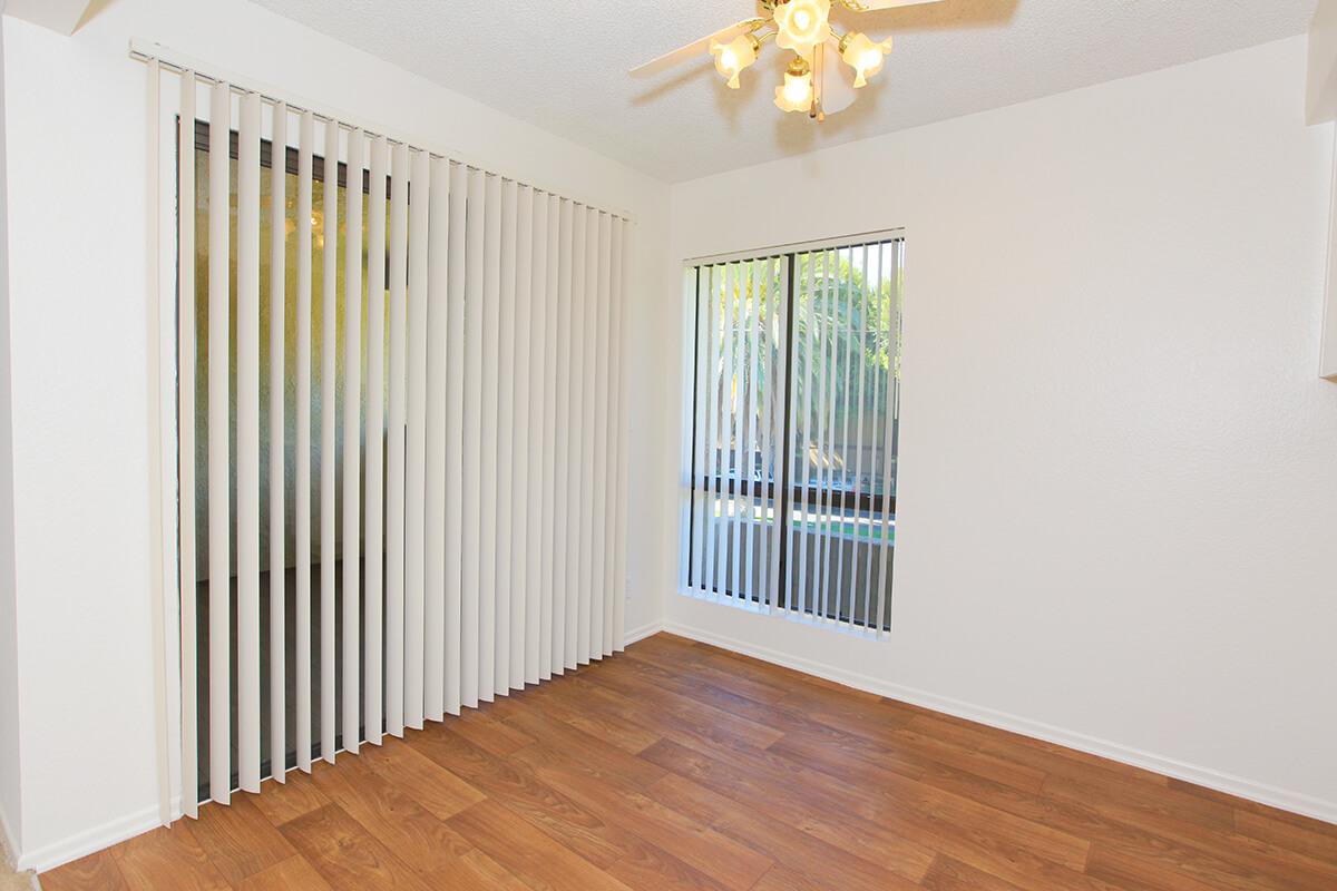 Bright, empty room with wooden flooring, featuring vertical blinds on large windows. A ceiling fan with light is visible, and part of the wall shows a small opening—possibly a pass-through to another room. Natural light illuminates the space, creating a welcoming atmosphere.