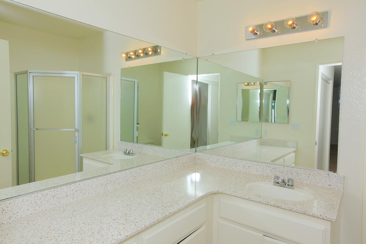 A modern bathroom featuring a spacious vanity with a white countertop and sink. Above the vanity, there are four light fixtures providing illumination. Large mirrors reflect the room, and a glass shower enclosure is visible in the background, along with a doorway leading to another area. The walls are painted a light color.