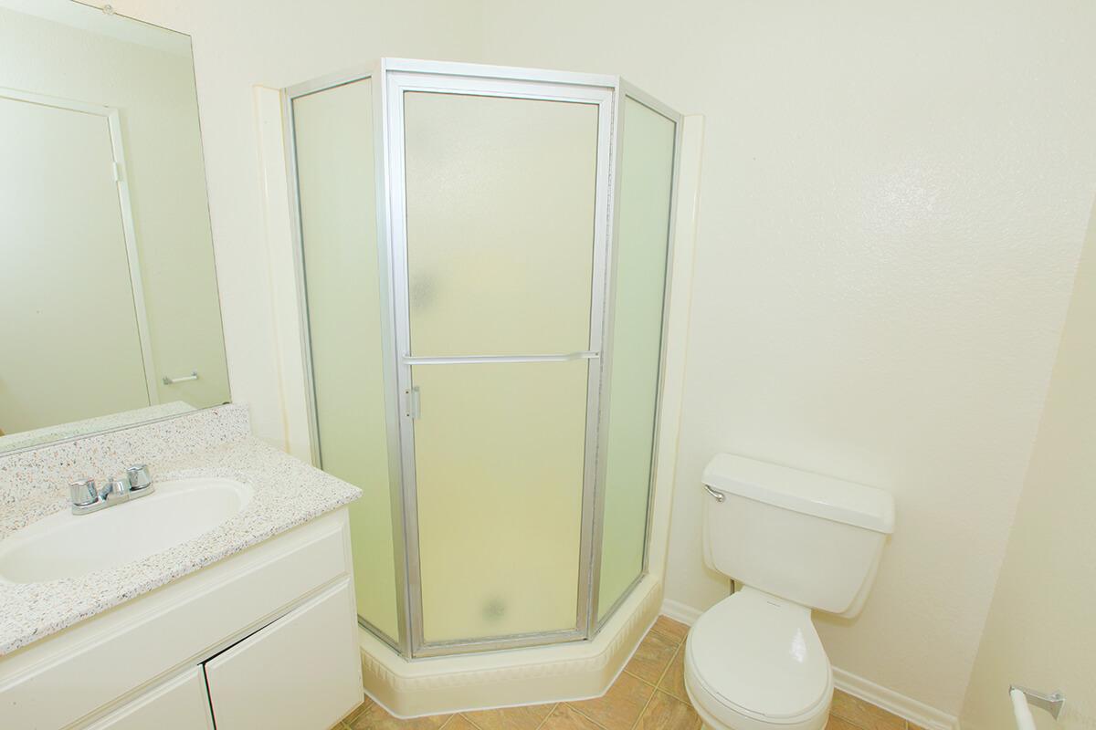 A clean, well-lit bathroom featuring a glass shower enclosure, toilet, and a sink with a countertop. The walls are painted a light color, and the flooring is tiled. A mirror is mounted above the sink, and the overall atmosphere is neat and tidy.