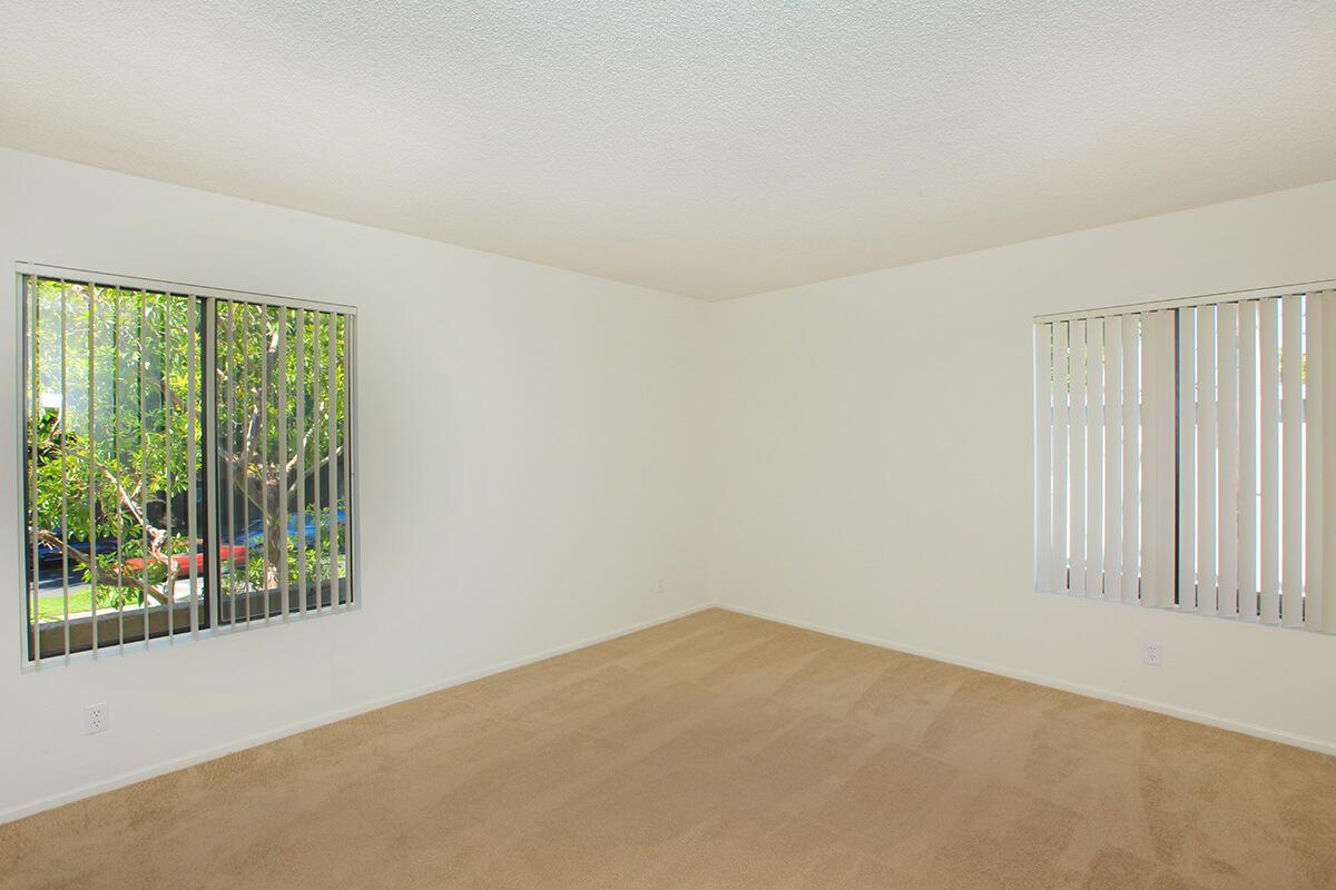 A spacious, unfurnished room with light beige carpet and white walls. There are two large windows with vertical blinds, allowing natural light to enter. Outside the windows, greenery is visible, creating a bright and airy atmosphere. The room is empty, suggesting it is ready for personal decoration.
