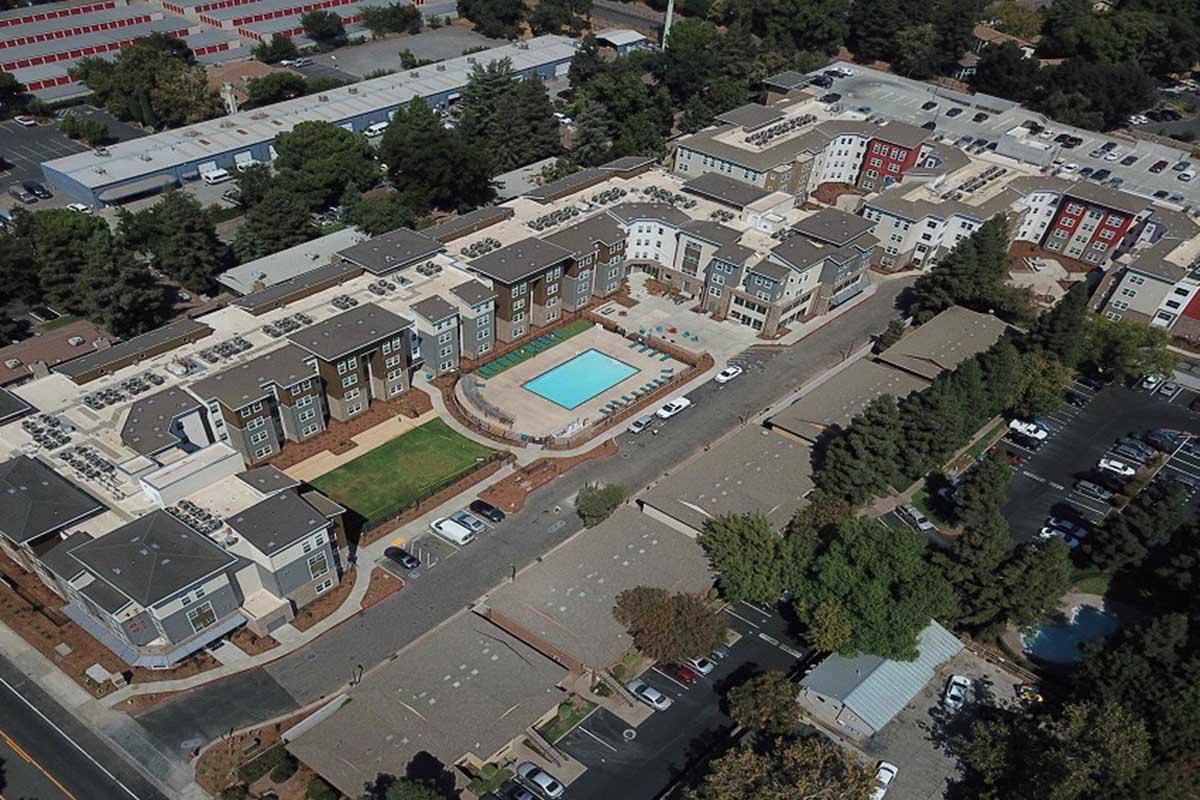 Aerial view of a modern apartment complex featuring multiple buildings surrounding a central swimming pool. The property is landscaped with trees and lawns, with parking lots visible nearby. The setting appears suburban, showcasing a blend of residential and green spaces.