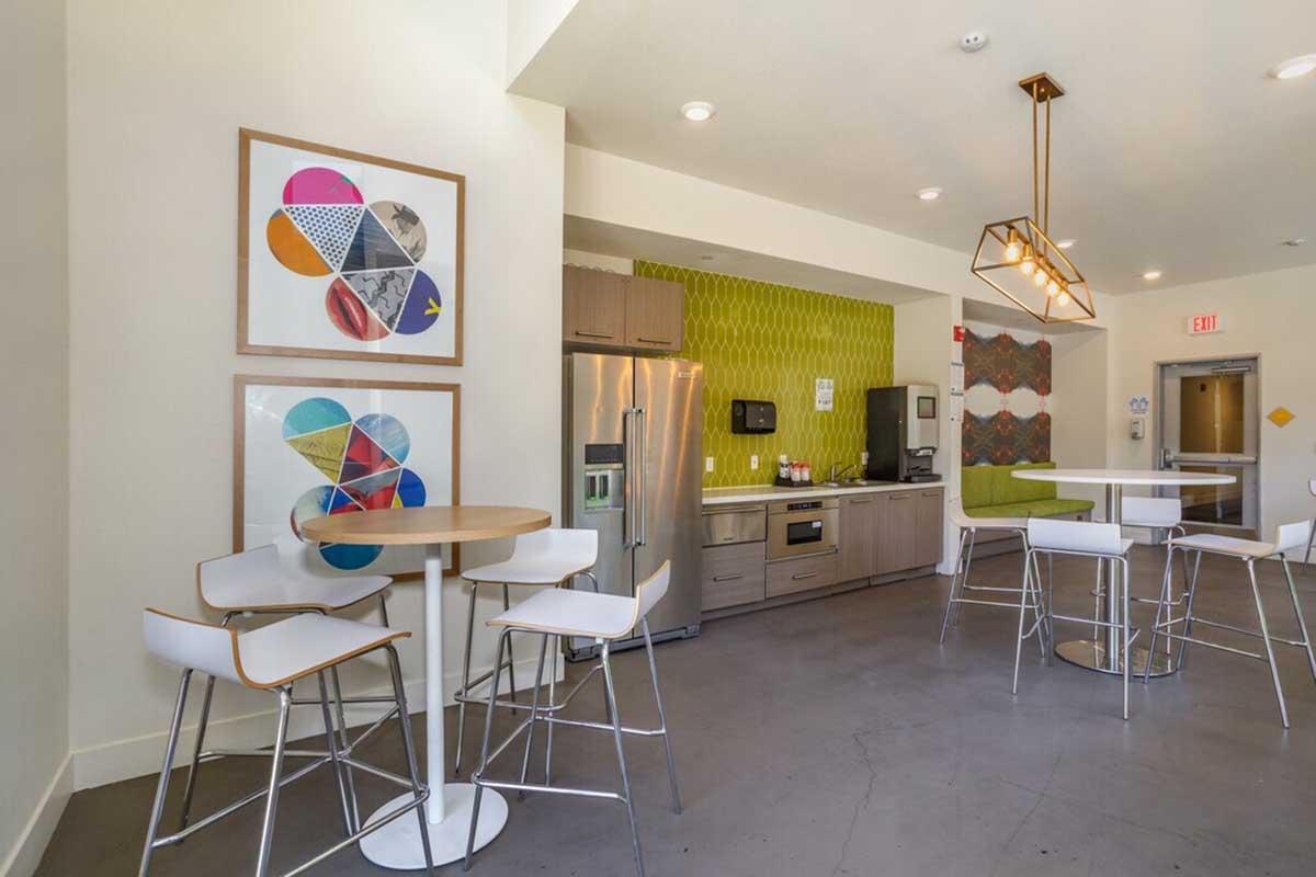 A modern kitchen area featuring a circular table with white chairs, a stainless steel refrigerator, and a green patterned wall. There are colorful framed artworks on the wall and a seating nook in the corner. The space is well-lit with a contemporary light fixture hanging above the table.