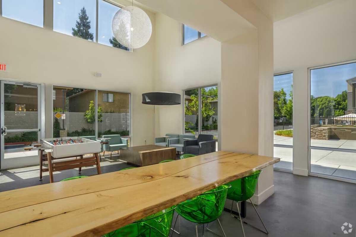 Bright modern lounge area featuring large windows, a wooden table with green chairs, and a foosball table. The space has minimalistic decor, with a cozy seating arrangement and access to an outdoor area visible through the windows.
