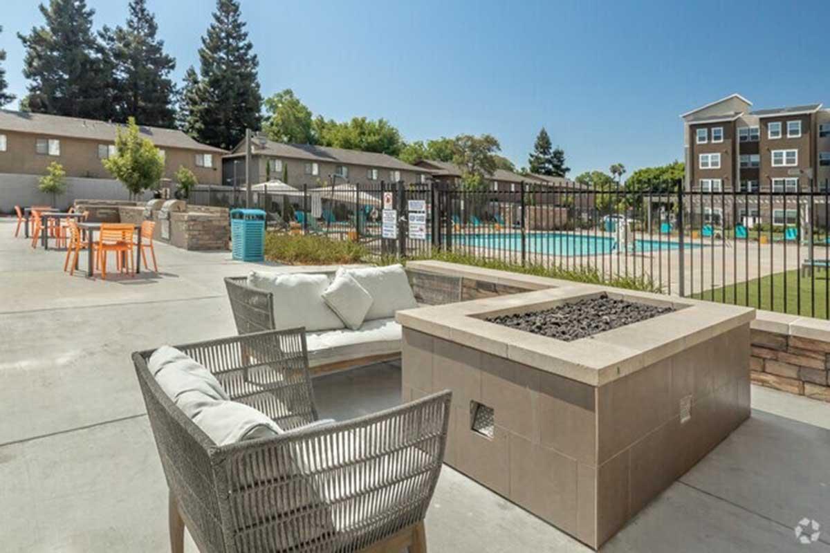 A modern outdoor area featuring a fire pit surrounded by comfortable seating. In the background, there is a swimming pool with lounge chairs and umbrellas, framed by green trees and apartments. The setting is bright and inviting, ideal for relaxation and social gatherings.