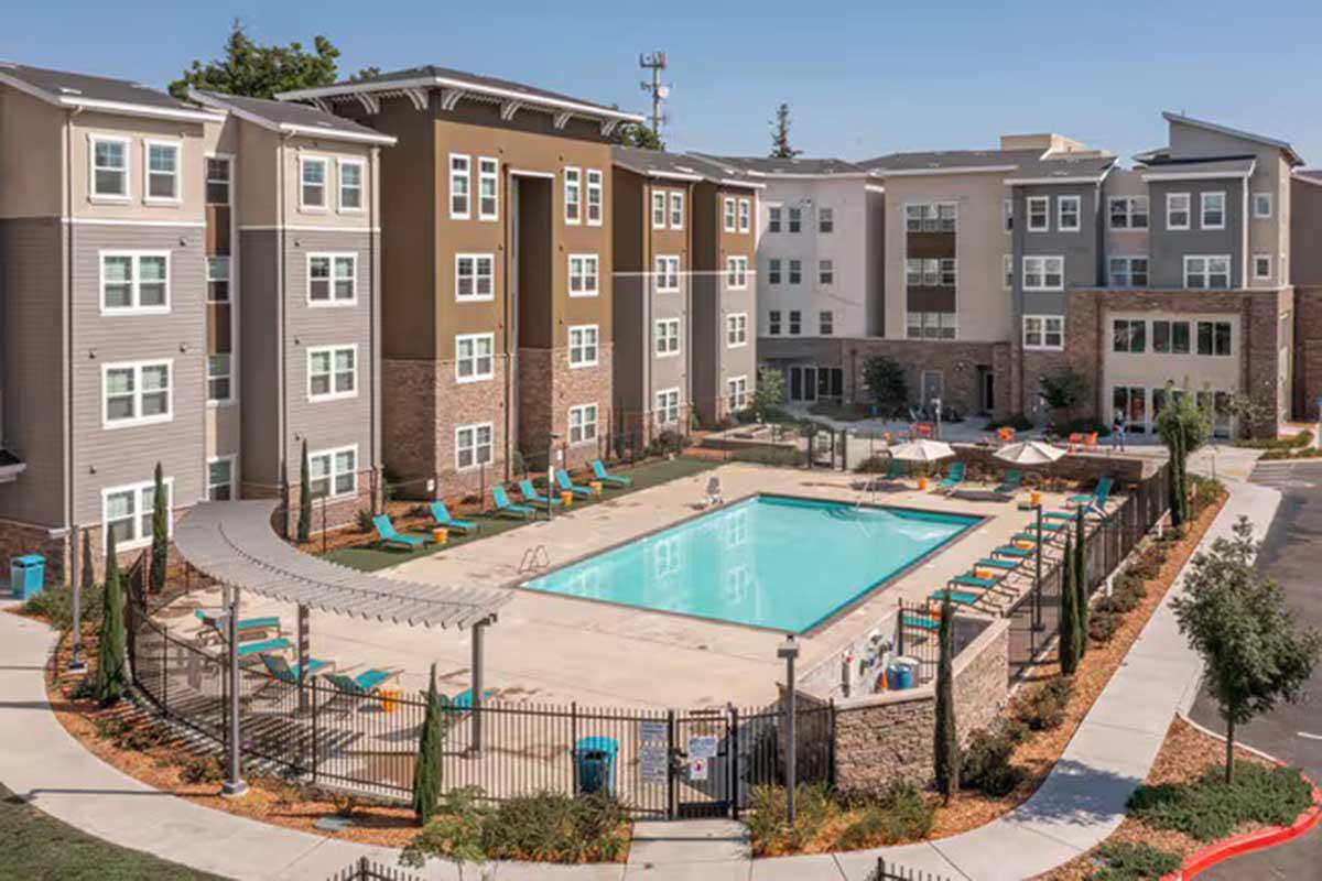 Aerial view of a modern apartment complex featuring a swimming pool surrounded by lounge chairs. The building has a mix of brown and beige exterior with balconies. Green landscaping and pathways lead to the pool area, creating an inviting outdoor space.