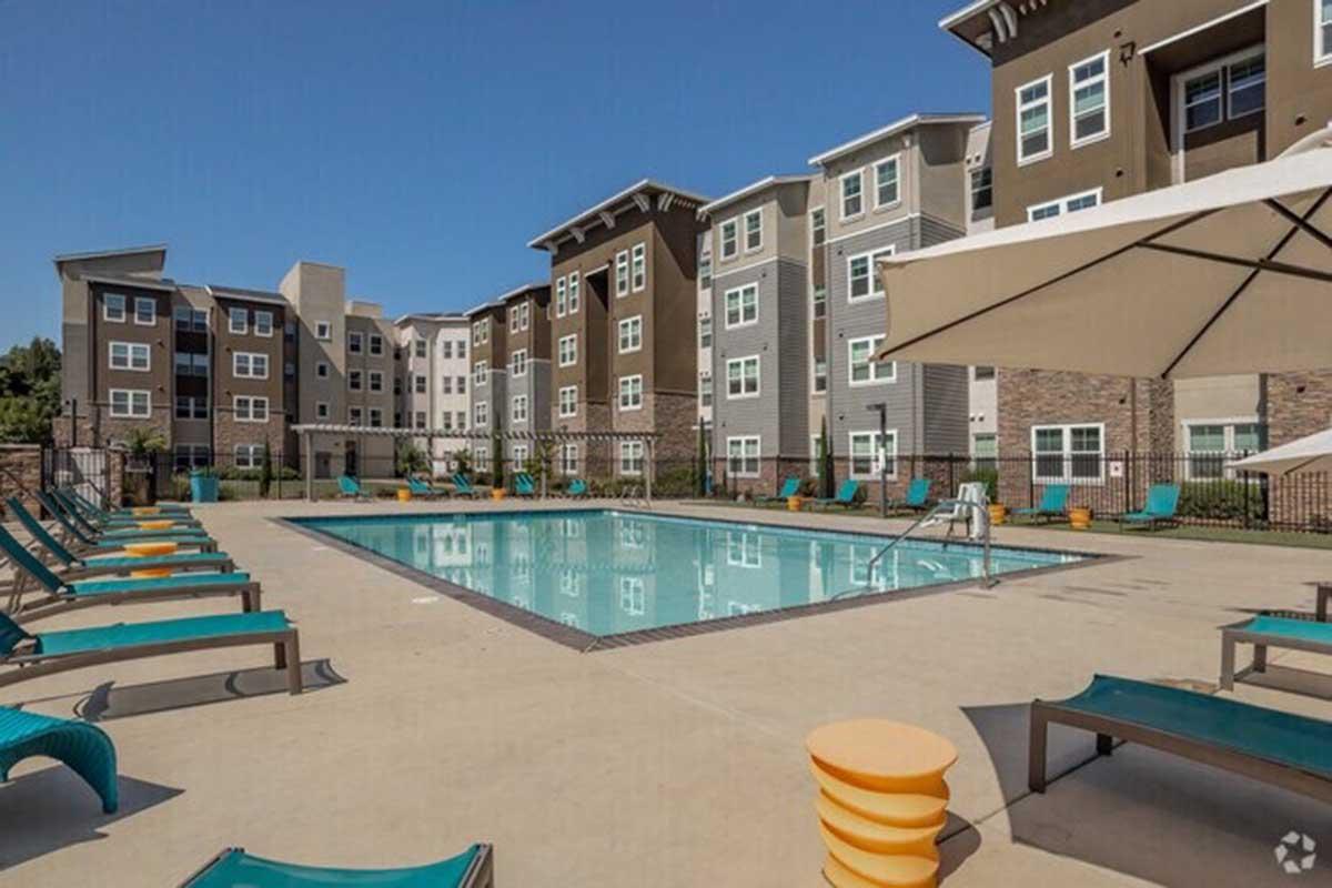 A vibrant outdoor pool area featuring a rectangular swimming pool surrounded by lounge chairs, umbrellas, and colorful furniture. Modern apartment buildings with various shades of brown and gray are in the background under a clear blue sky.
