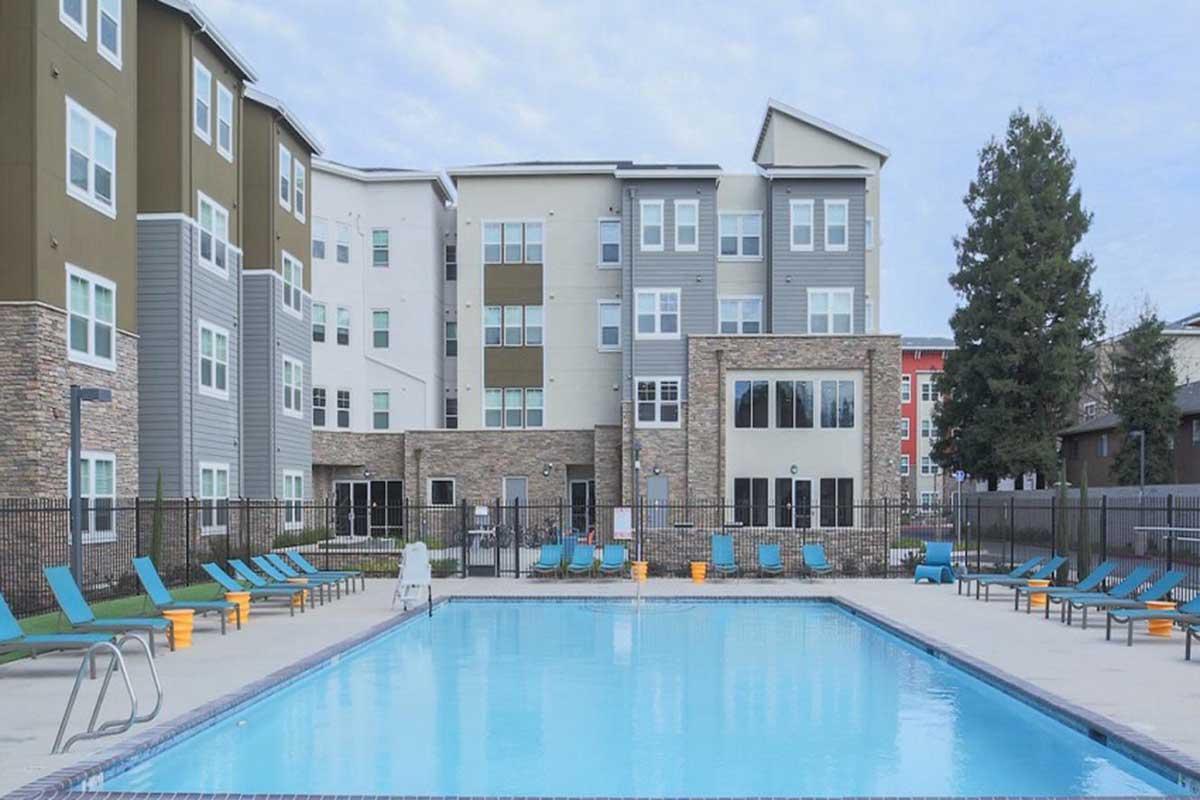 A spacious outdoor swimming pool surrounded by lounge chairs, located in a residential complex. The pool area features a modern building in the background with green, brown, and gray architecture. Several trees are also visible, adding to the inviting atmosphere of the area.