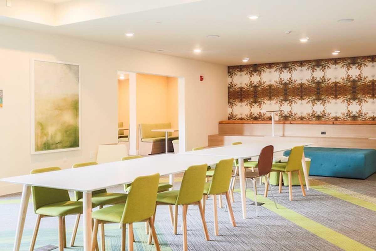 A modern meeting room featuring a long white table surrounded by green chairs. There is a patterned accent wall and soft lighting. The space includes cozy seating areas in the background, creating an inviting atmosphere for collaboration and gatherings.