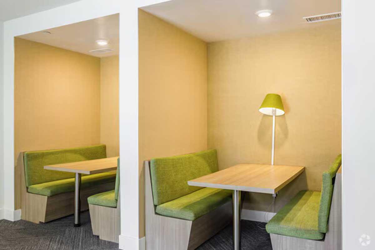 Cozy seating area with two enclosed booths featuring green upholstery and wooden tables. Each booth is illuminated by a modern lamp with a green shade, set against a light yellow wall, creating a comfortable and inviting atmosphere.