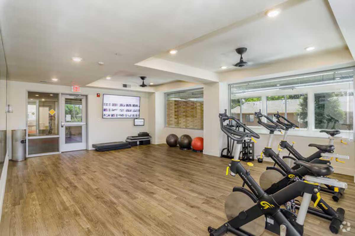 A well-lit fitness room featuring several exercise bikes, yoga mats, and exercise balls. Large windows provide natural light, while ceiling fans help circulate air. The space is clean and modern, designed for workouts and physical activity.
