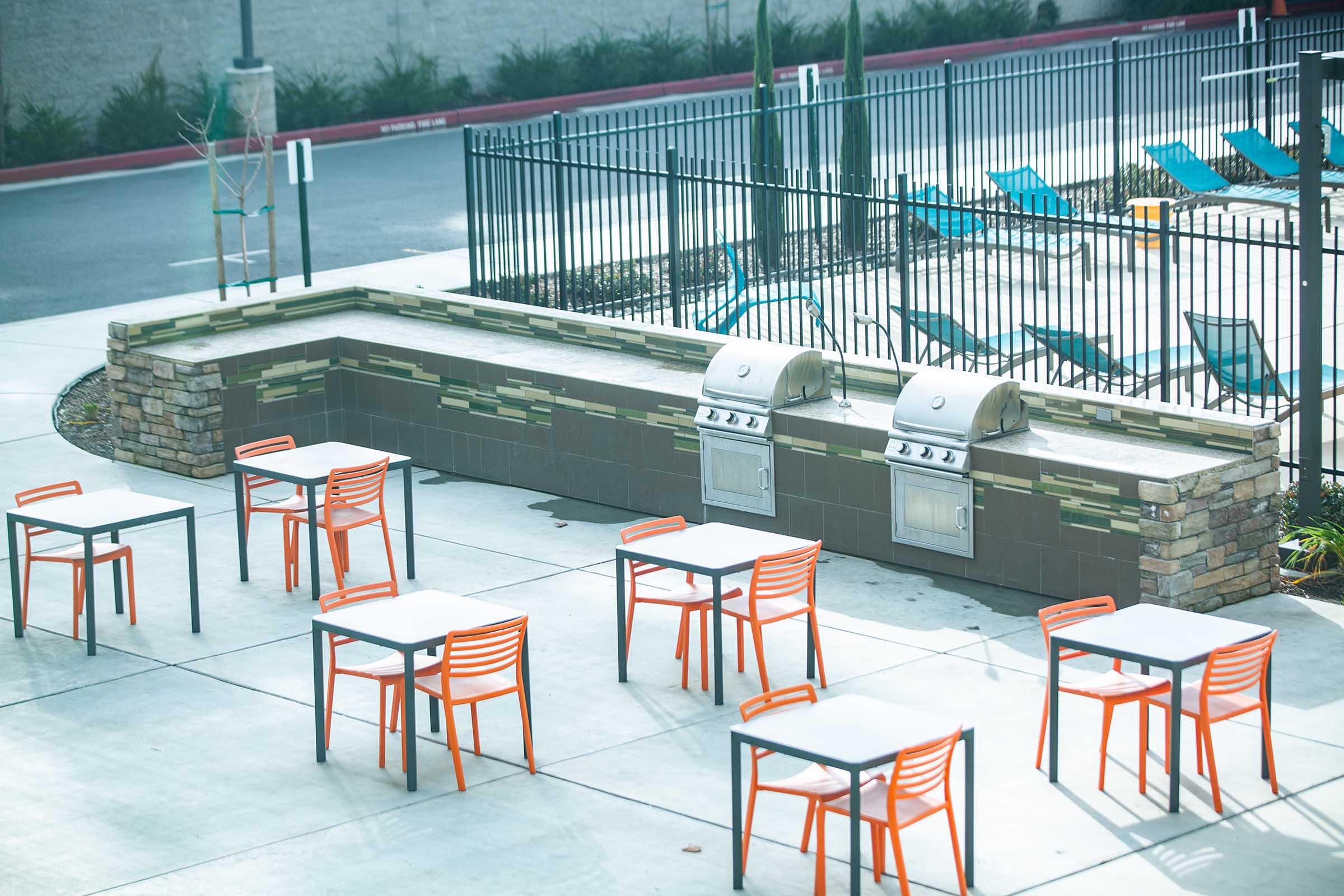 Outdoor patio area featuring a stone barbecue grill station, two grills, and several empty tables with orange chairs. A fenced pool area with lounge chairs is visible in the background. The setting is bright and inviting, suitable for gatherings or barbecue events.