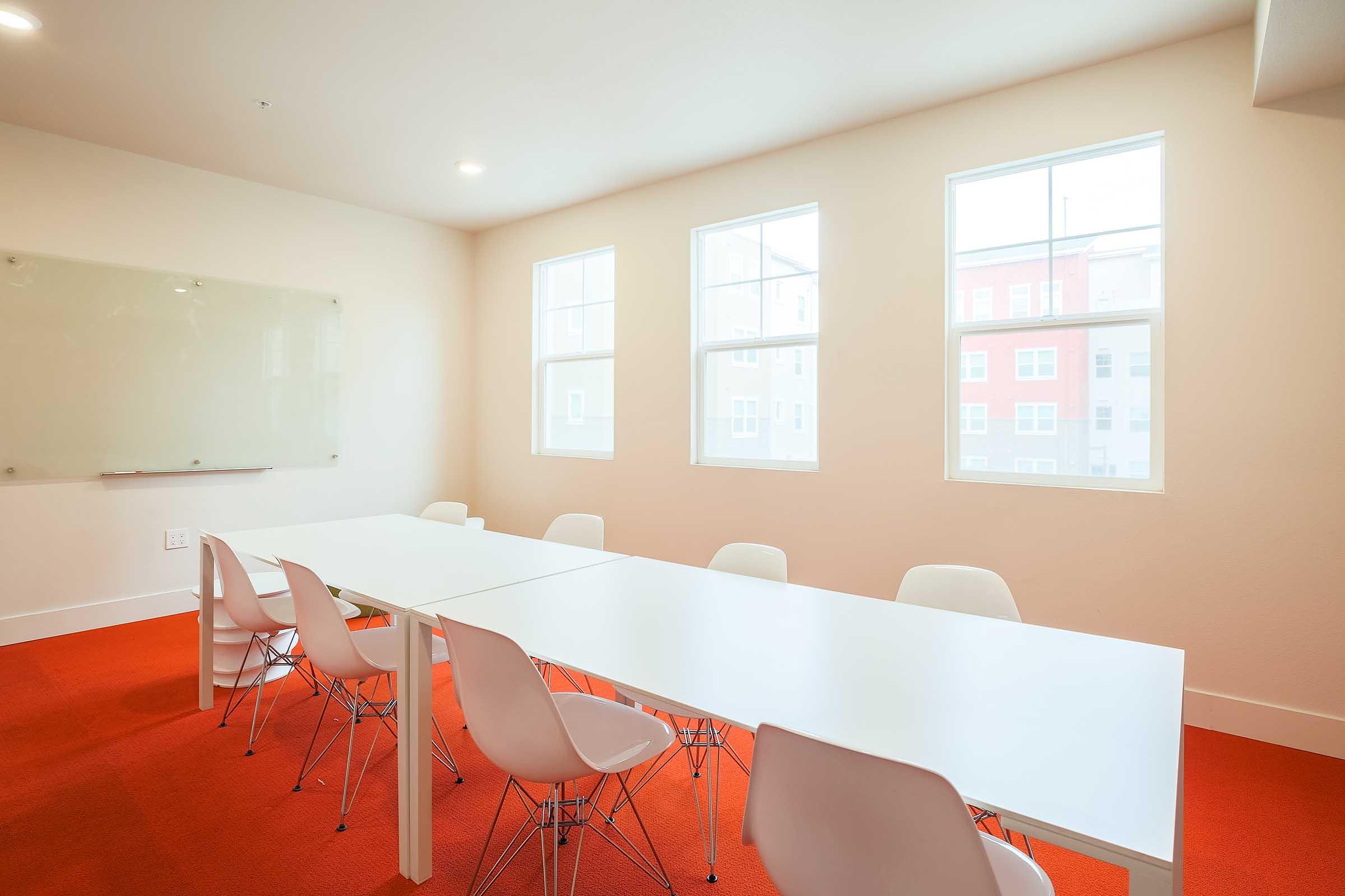 A modern conference room featuring a long white table surrounded by several white chairs. The room has three large windows allowing natural light to enter, and a glass whiteboard is mounted on the wall. The floor is covered with bright red carpeting, creating a vibrant atmosphere.