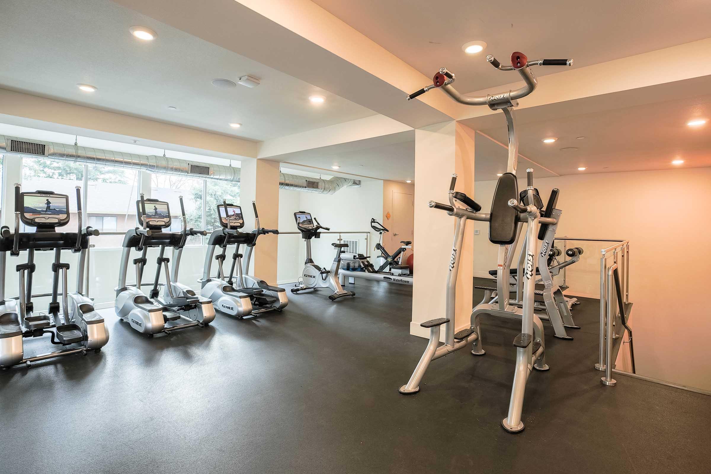 A modern gym interior featuring multiple elliptical machines lined up against a wall, with an open area for workouts. Bright lighting and large windows create a welcoming atmosphere, while a stair-step machine is positioned nearby, showcasing an organized fitness space.