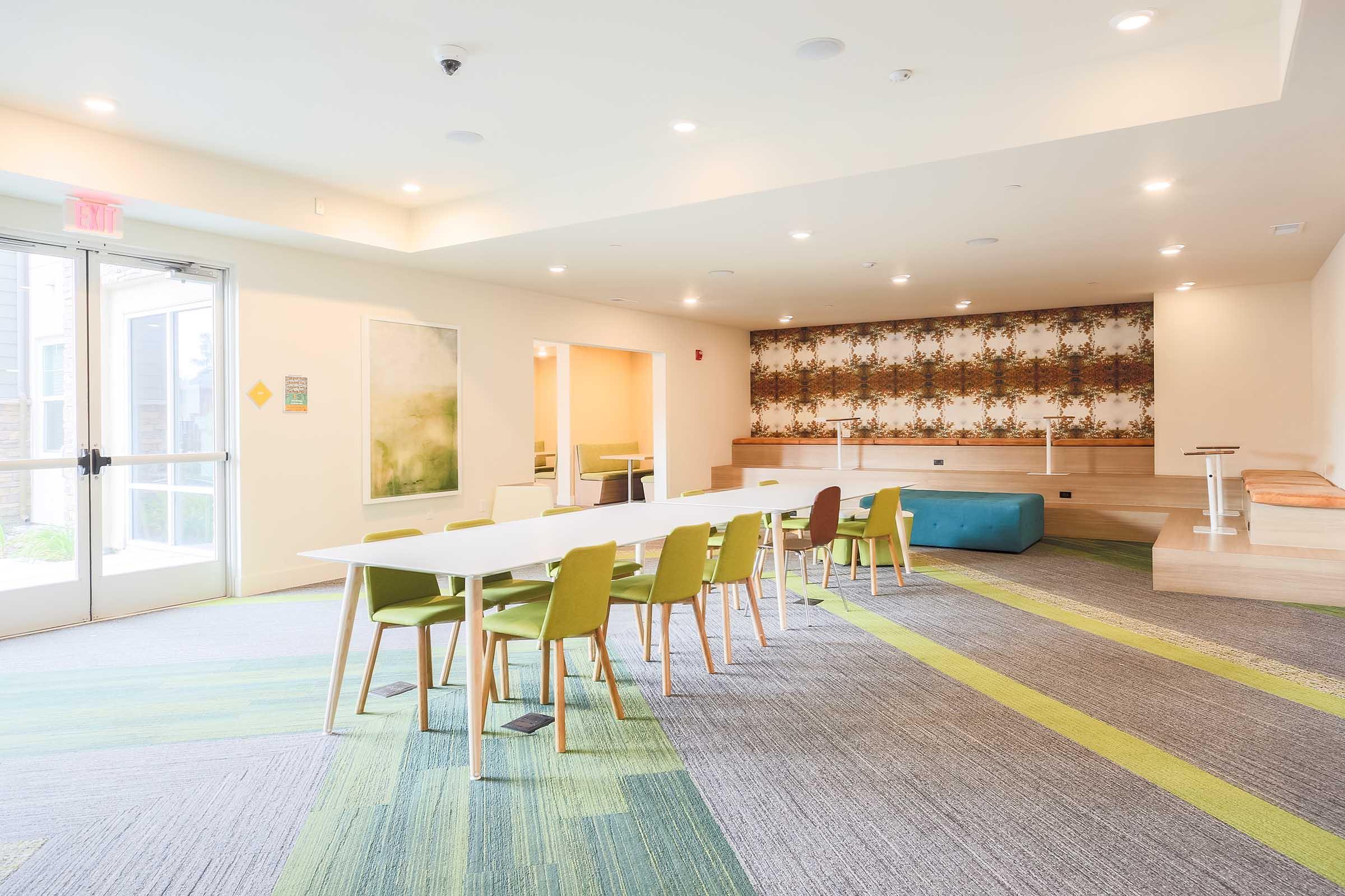 A brightly lit modern meeting room featuring a long white table surrounded by green chairs. The space includes wood accents, patterned wallpaper on one wall, and a blue seating area. Large windows provide natural light, creating an inviting atmosphere for collaboration or gatherings.