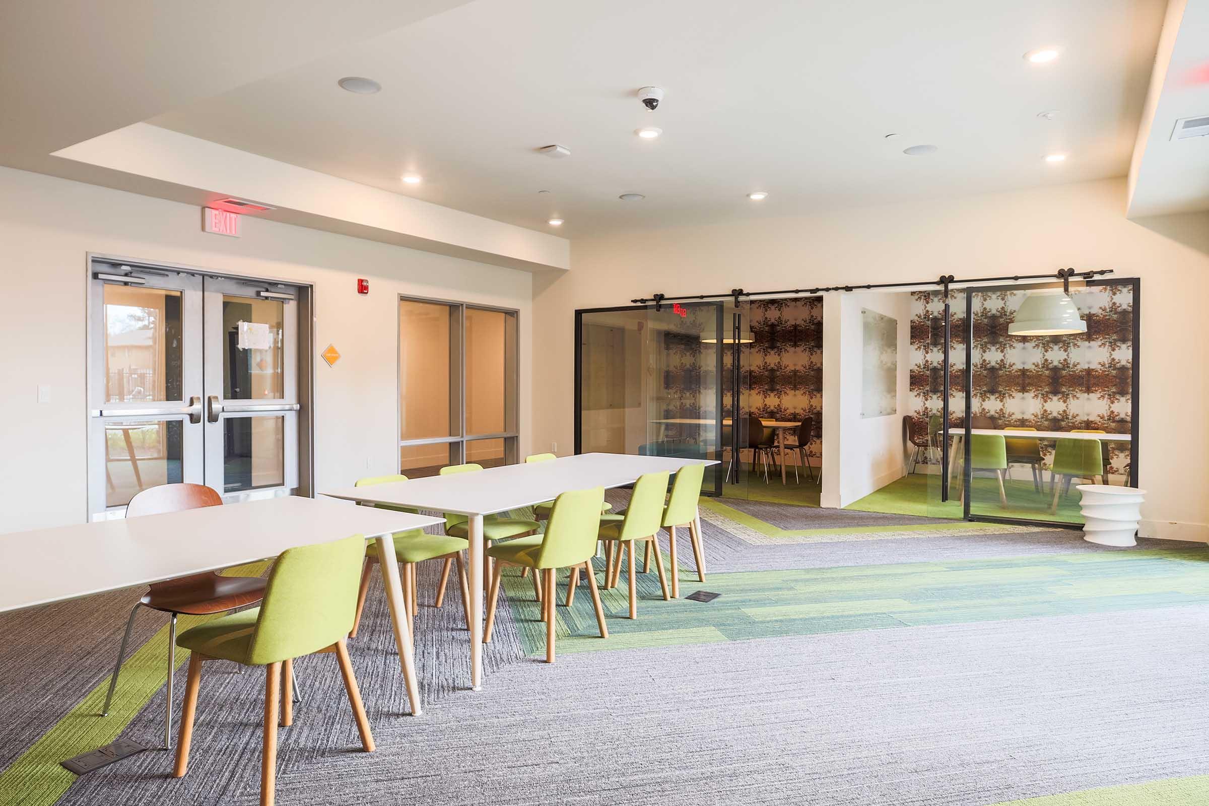 A modern, well-lit office or meeting space featuring a long white table with green and wooden chairs. In the background, there are glass doors leading to a private area with patterned walls, and additional seating can be seen. The floor is covered in green and gray carpeting, creating a contemporary atmosphere.