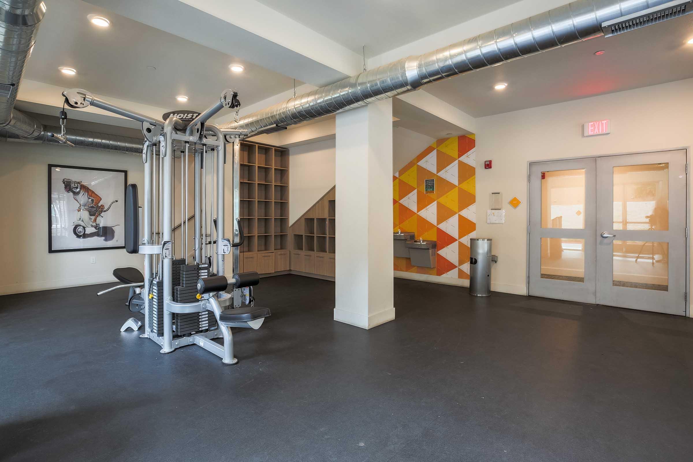 A modern gym interior featuring a multi-functional weight machine, shelving unit, and a colorful geometric wall. There are two doors leading to other areas, an exit sign, and a water fountain visible in the space. The overall design is sleek and well-lit.