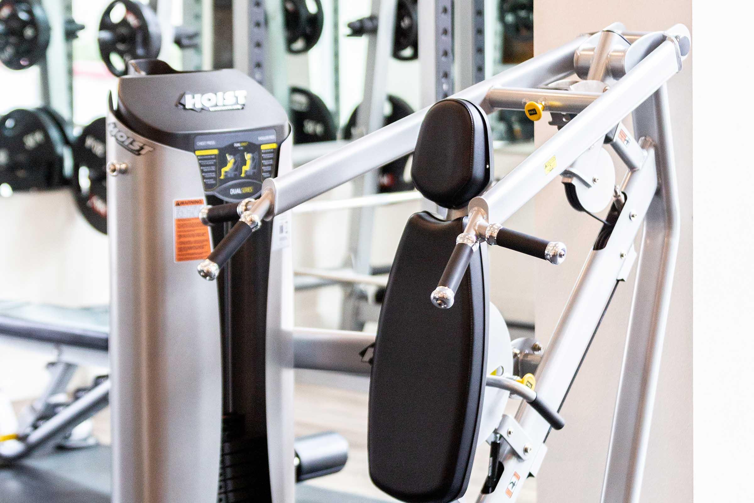 A fitness machine designed for strength training, featuring adjustable seats and handlebars. It is located in a gym environment with weights and exercise equipment visible in the background. The machine is made of metal and has a user-friendly display panel.