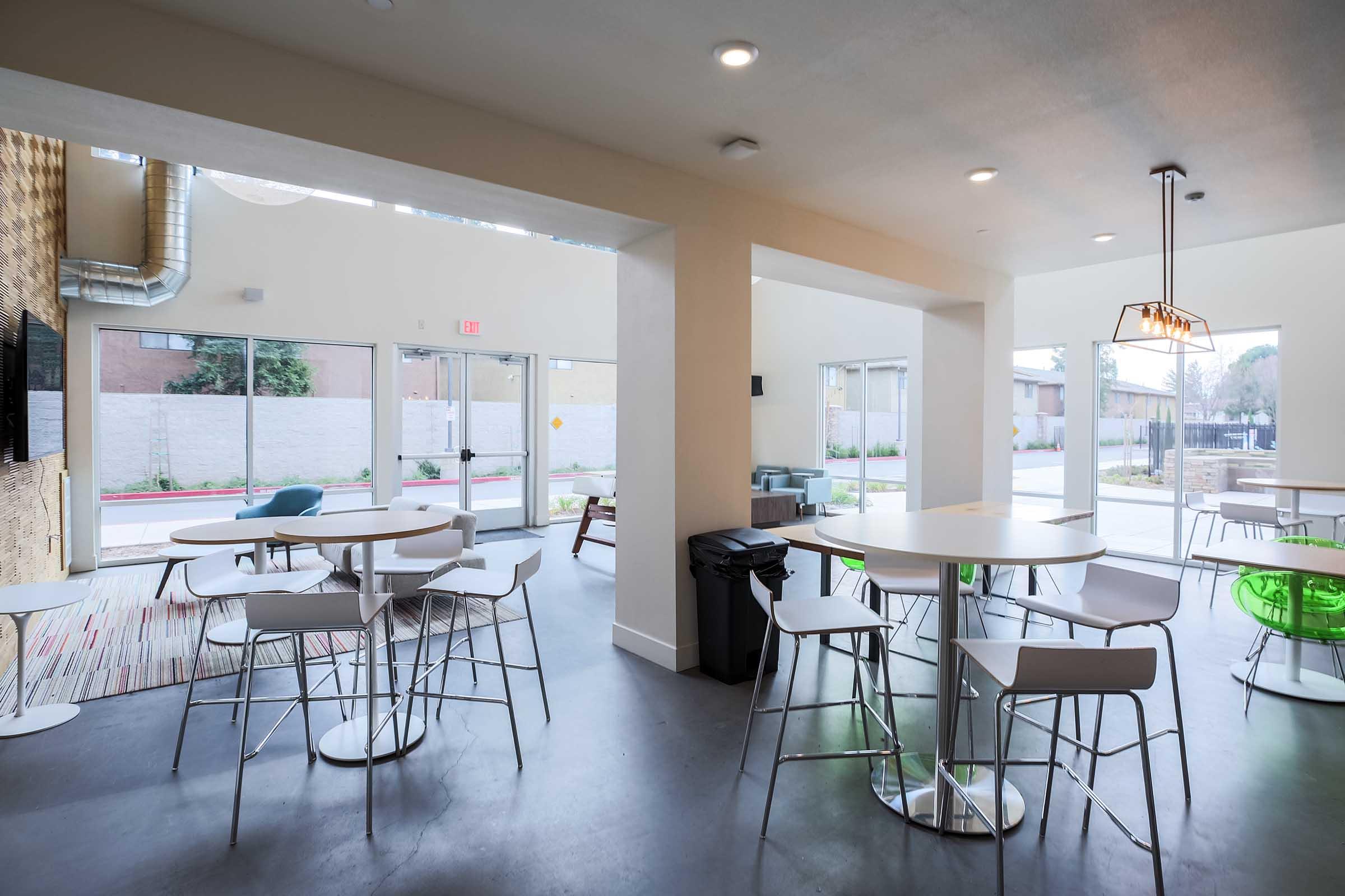 Indoor communal space featuring several round tables with white chairs, a few green stools, and large windows allowing natural light. A trash can is visible, and there are decorative elements in the background. The setting appears modern and inviting, suitable for gatherings or casual meetings.