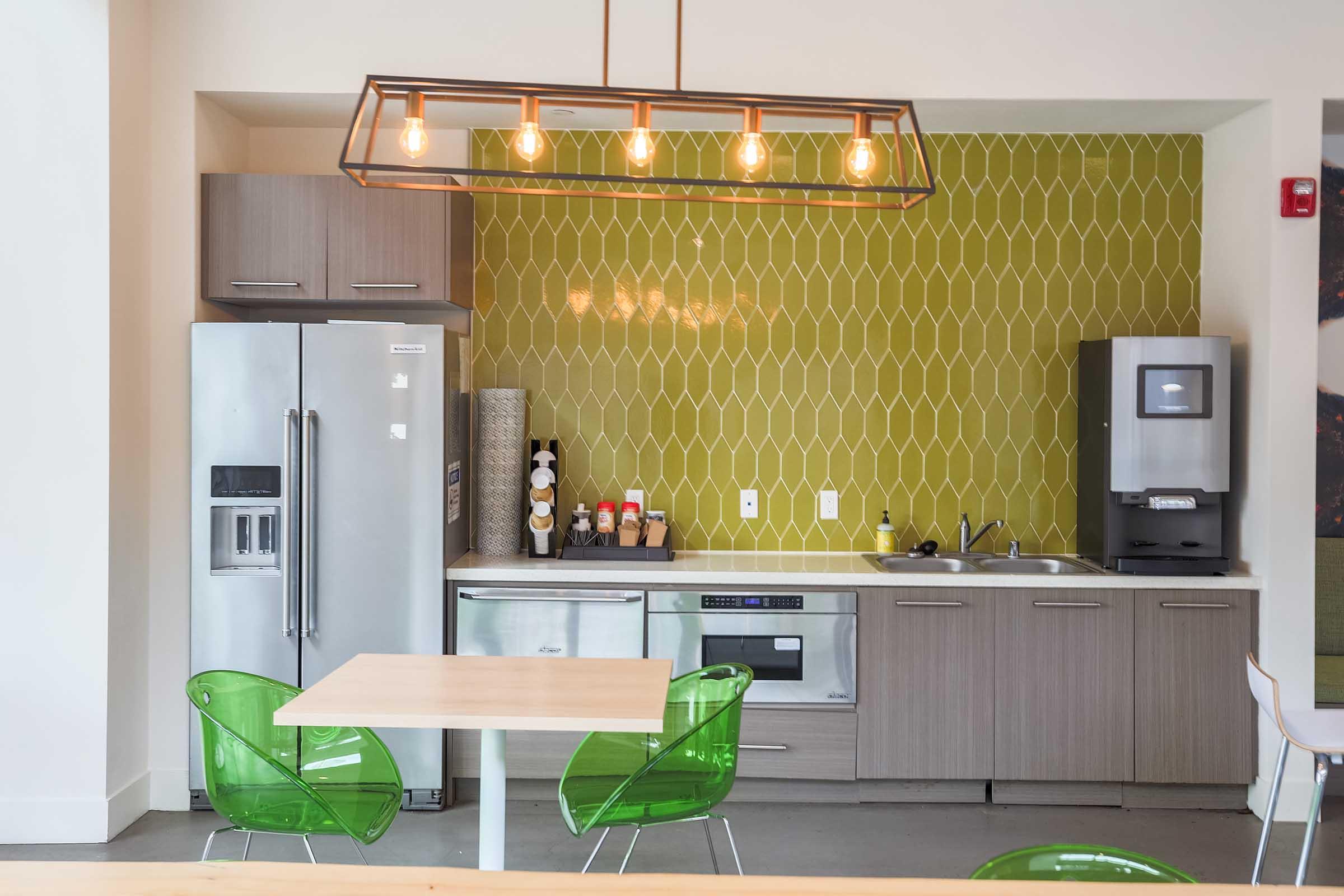 Modern kitchen featuring stainless steel appliances, a green tiled backsplash, and a wooden table with transparent green chairs. The lighting includes a stylish pendant fixture with exposed bulbs, creating a contemporary and inviting atmosphere.