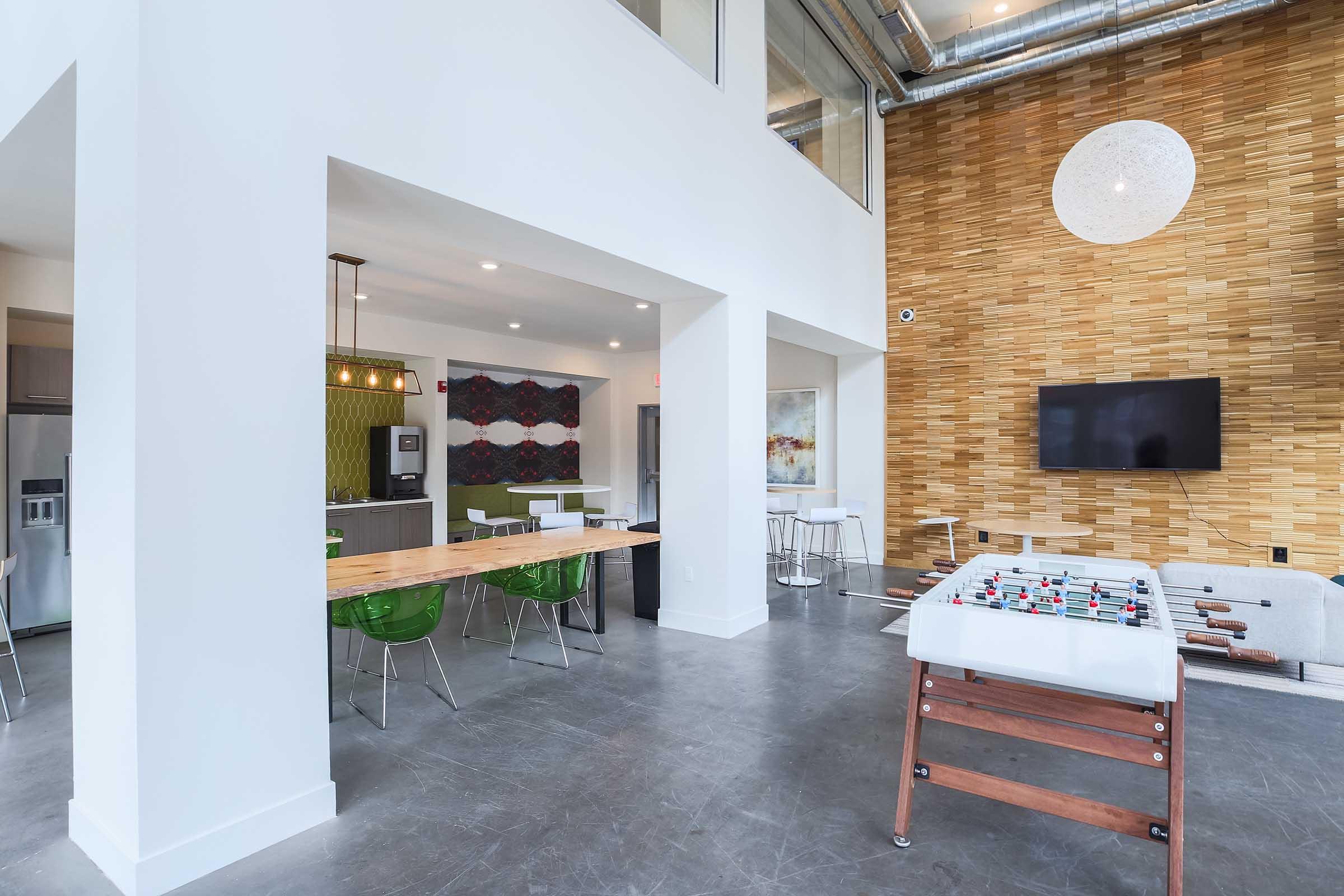 Modern interior of an open-concept space featuring a foosball table, a dining area with green chairs, a kitchenette in the background, and a large wall-mounted TV. The decor includes a textured wooden wall and contemporary lighting. Bright and airy atmosphere with large windows.