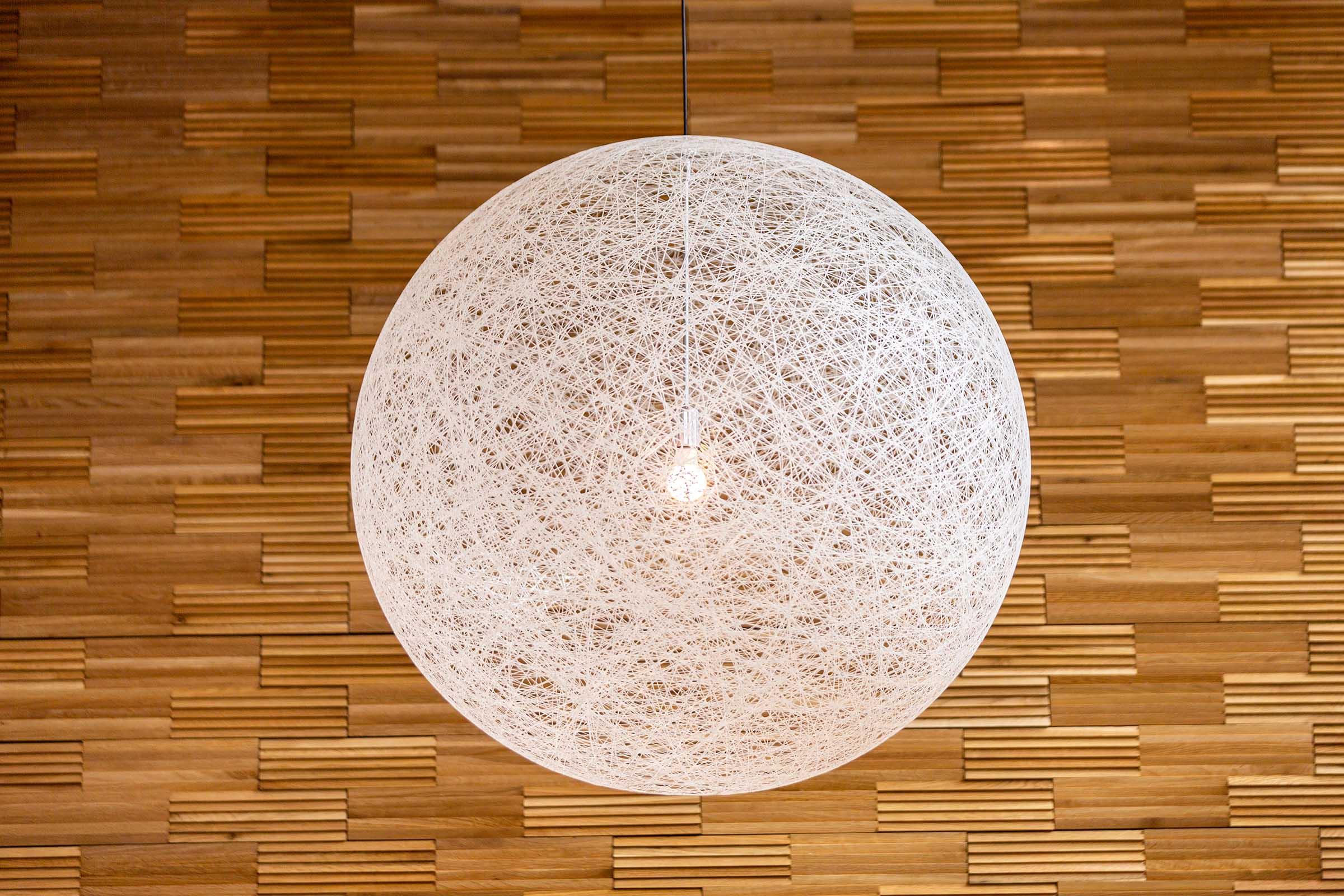 A large white spherical pendant light made of intertwined string or wire, illuminated from within. The light hangs against a backdrop of wooden wall panels featuring a textured, modern design.