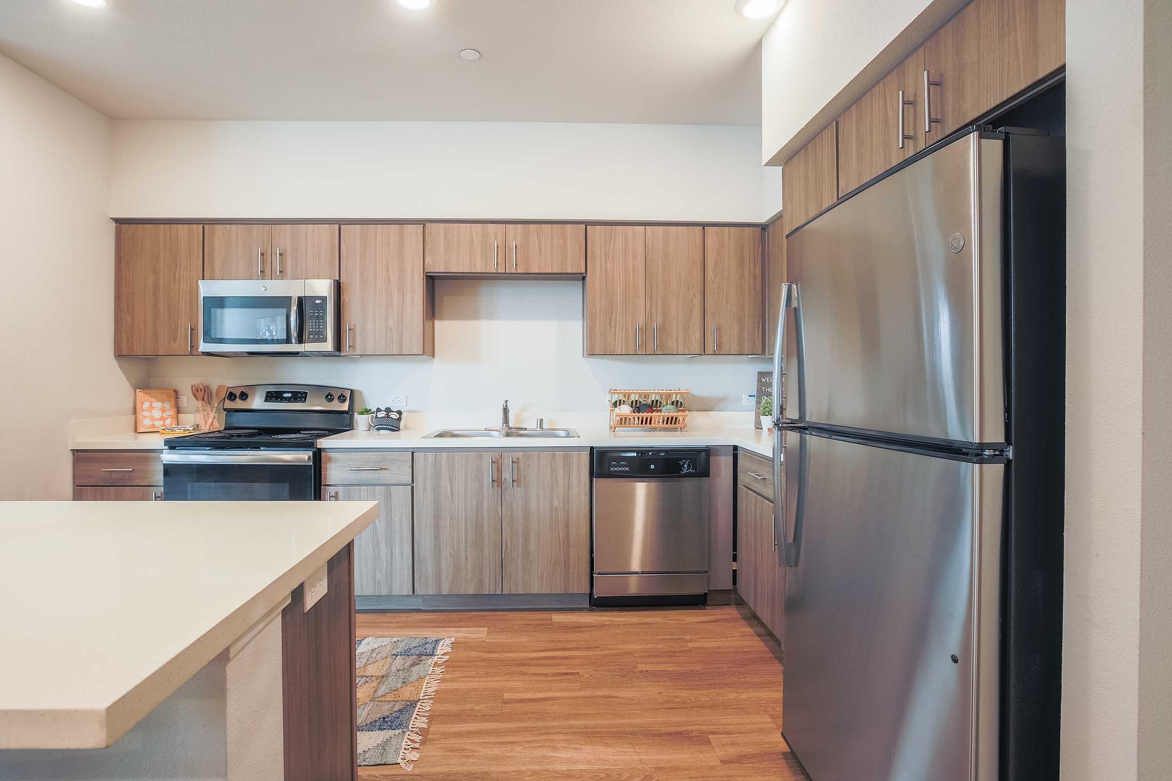 Modern kitchen with wooden cabinetry, stainless steel appliances including a refrigerator, oven, and dishwasher. A large island with a white countertop is in the foreground. Light wood flooring complements the contemporary design. Well-lit space with a clean and organized layout.