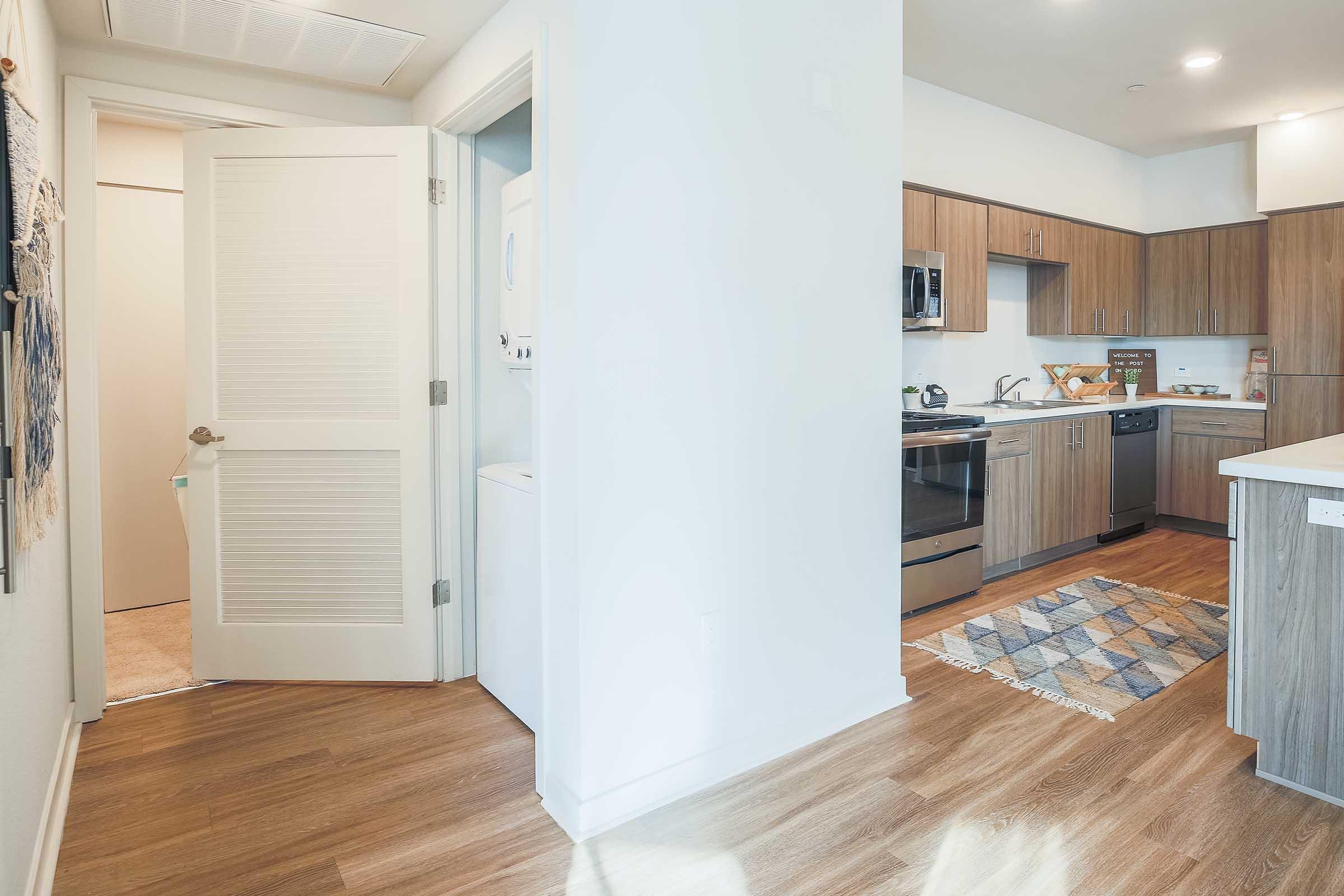 A modern kitchen with wooden cabinets and stainless steel appliances, including a stove and dishwasher. To the left, a beige door leads to another room. A washing machine is visible in a closet. The floor is light wood, and a patterned rug adds color to the space, creating a welcoming atmosphere.