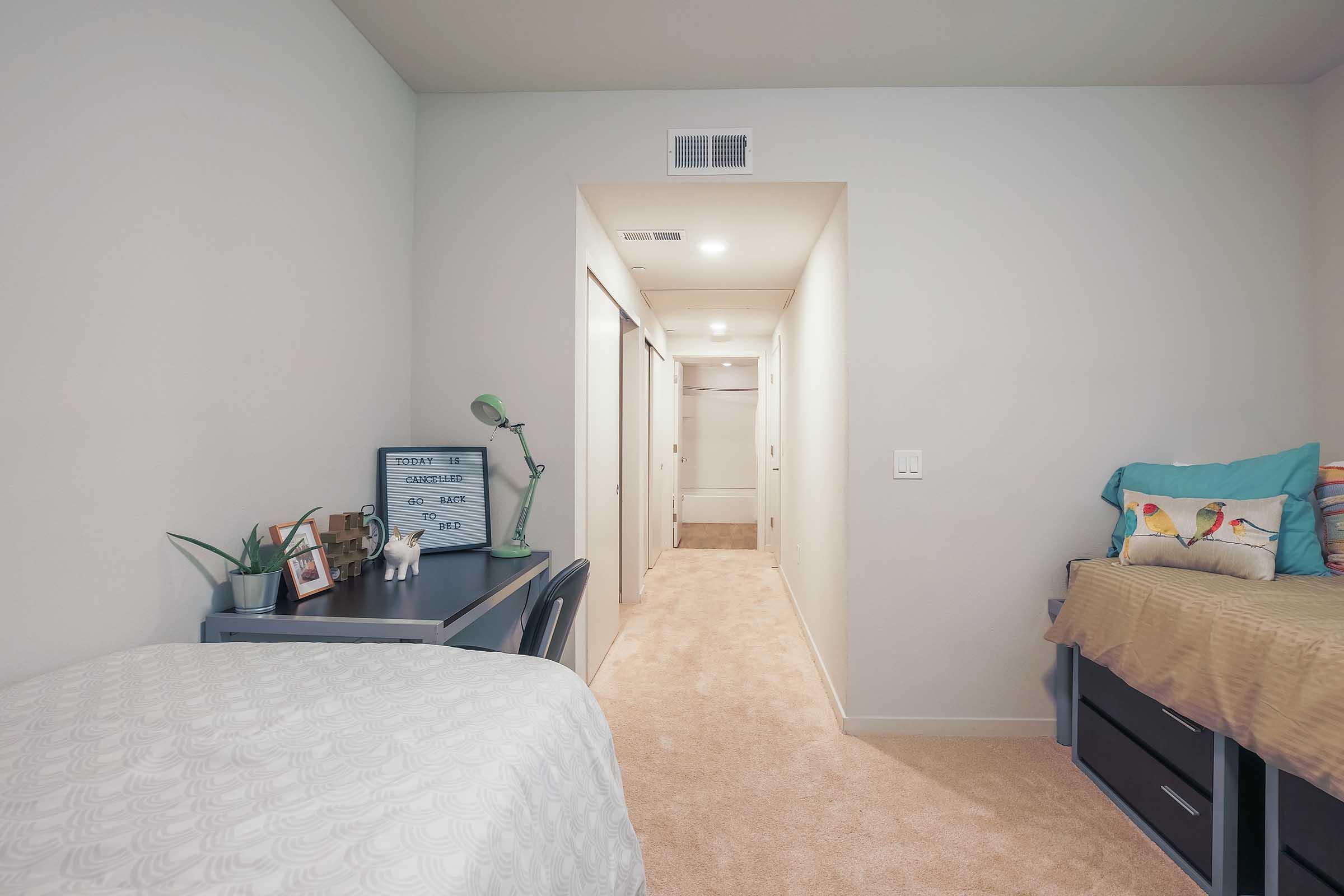 A bedroom featuring two beds, one with colorful pillows, and a small desk with a lamp and plant. A hallway leads to an area with closet doors, and the walls are painted a light color. The carpet is beige, creating a cozy atmosphere.