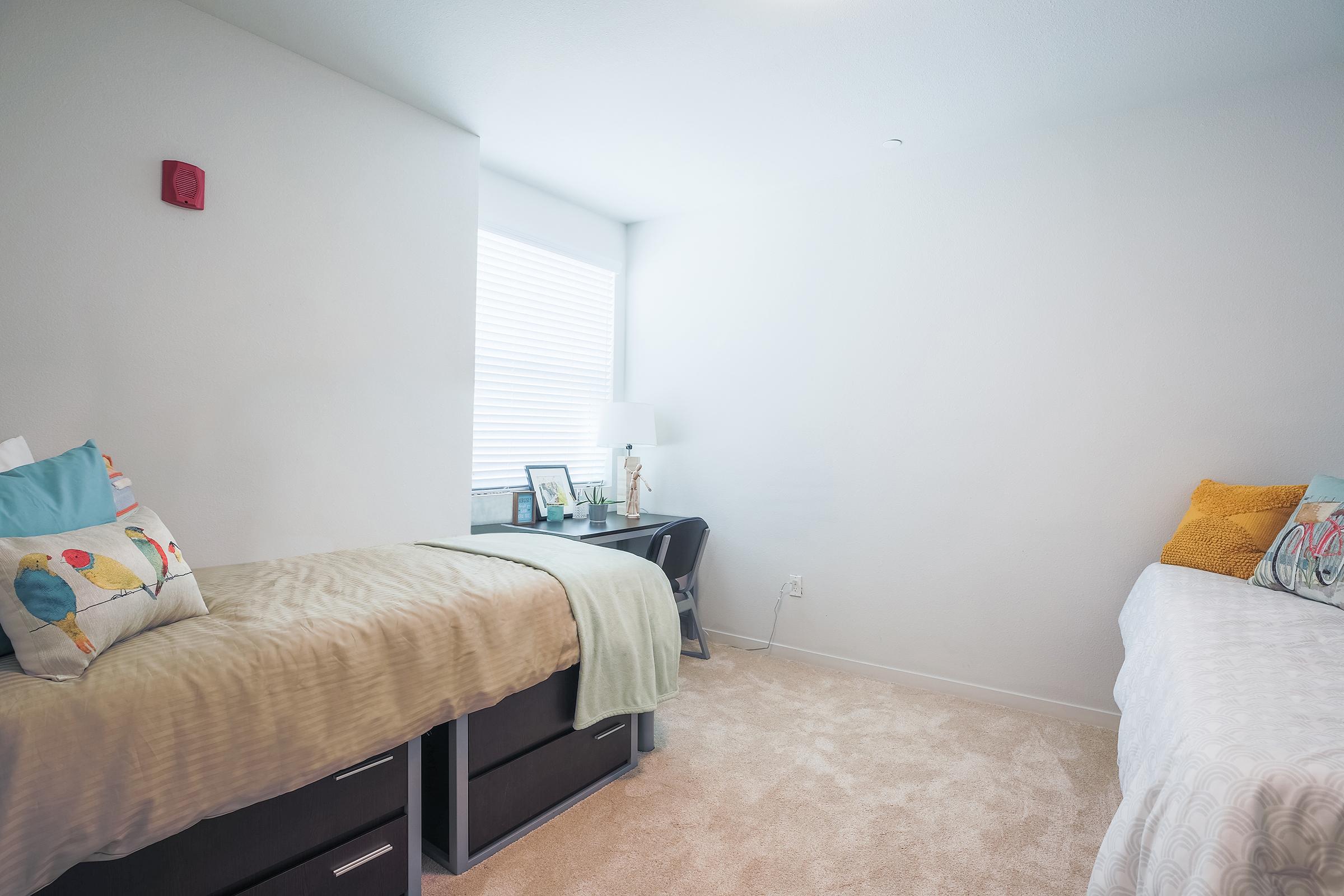 A tidy bedroom featuring two twin beds with colorful bedding, a small desk with a lamp and framed photos, and a window with blinds. The room has light-colored walls and carpet, creating a bright and inviting atmosphere.