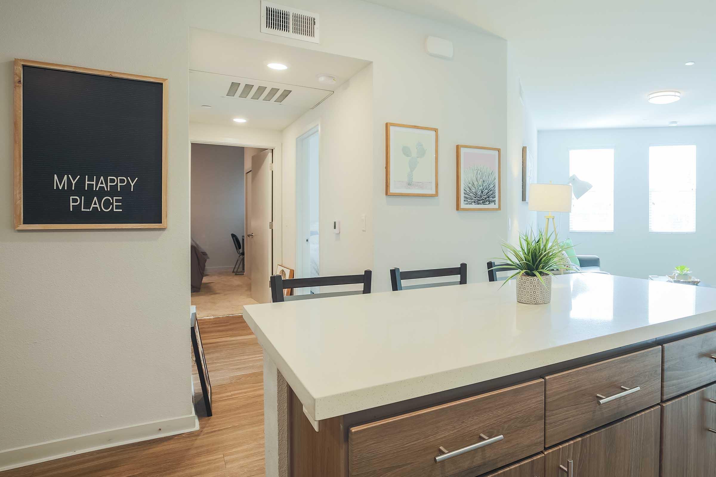 A modern kitchen with a light-colored countertop, dark wood cabinets, and black barstools. A decorative chalkboard on the wall reads "MY HAPPY PLACE." In the background, a cozy living area is visible, with natural light coming through large windows. Plants and decor enhance the inviting atmosphere.