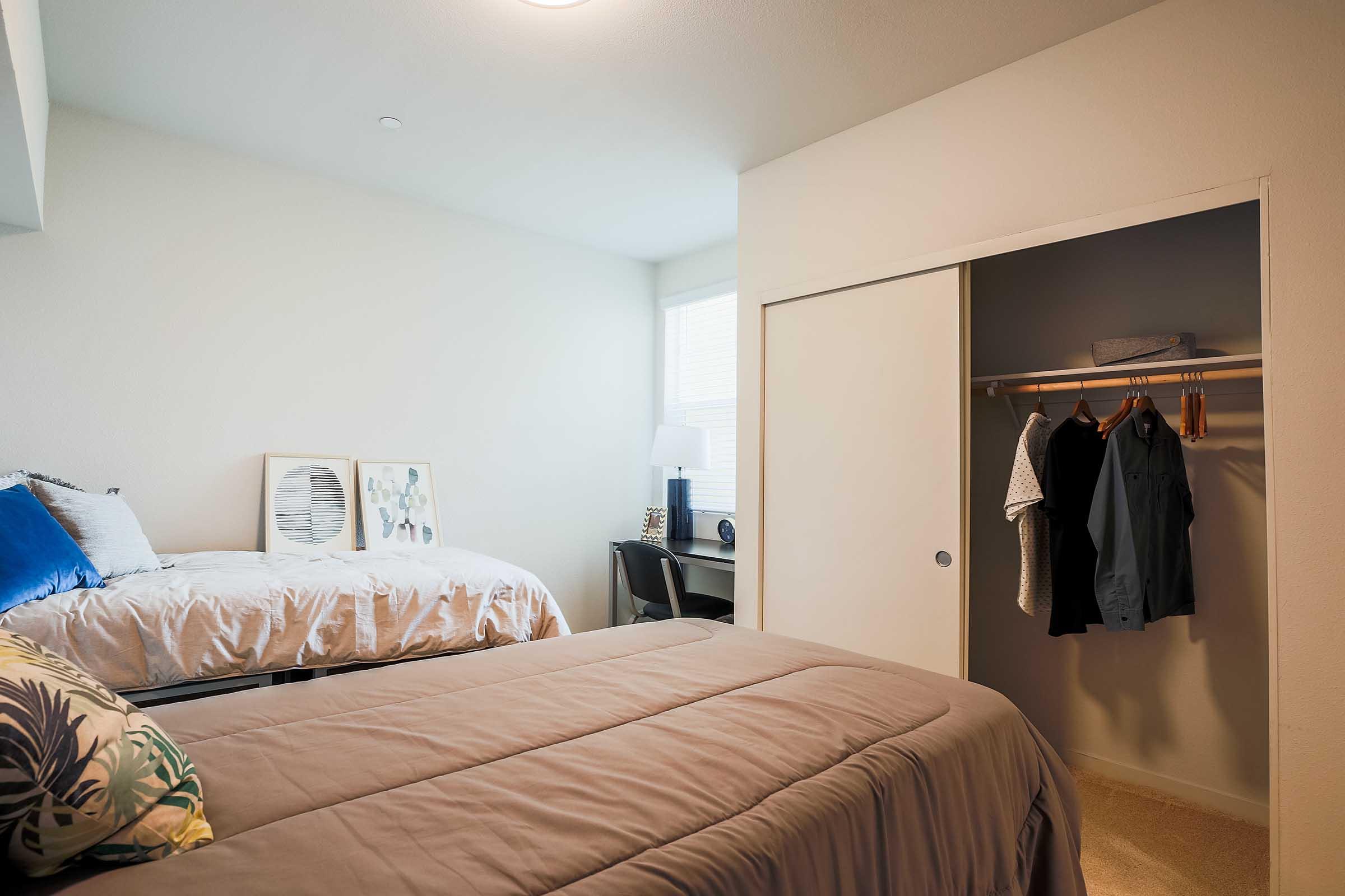 Two neatly made beds in a light, minimalist bedroom. One bed is against the wall with decorative pillows, while the other is slightly ajar. There's a small desk with a lamp near a window, and a closet showcasing hanging clothes. The walls are painted a soft color, enhancing the airy feel of the room.