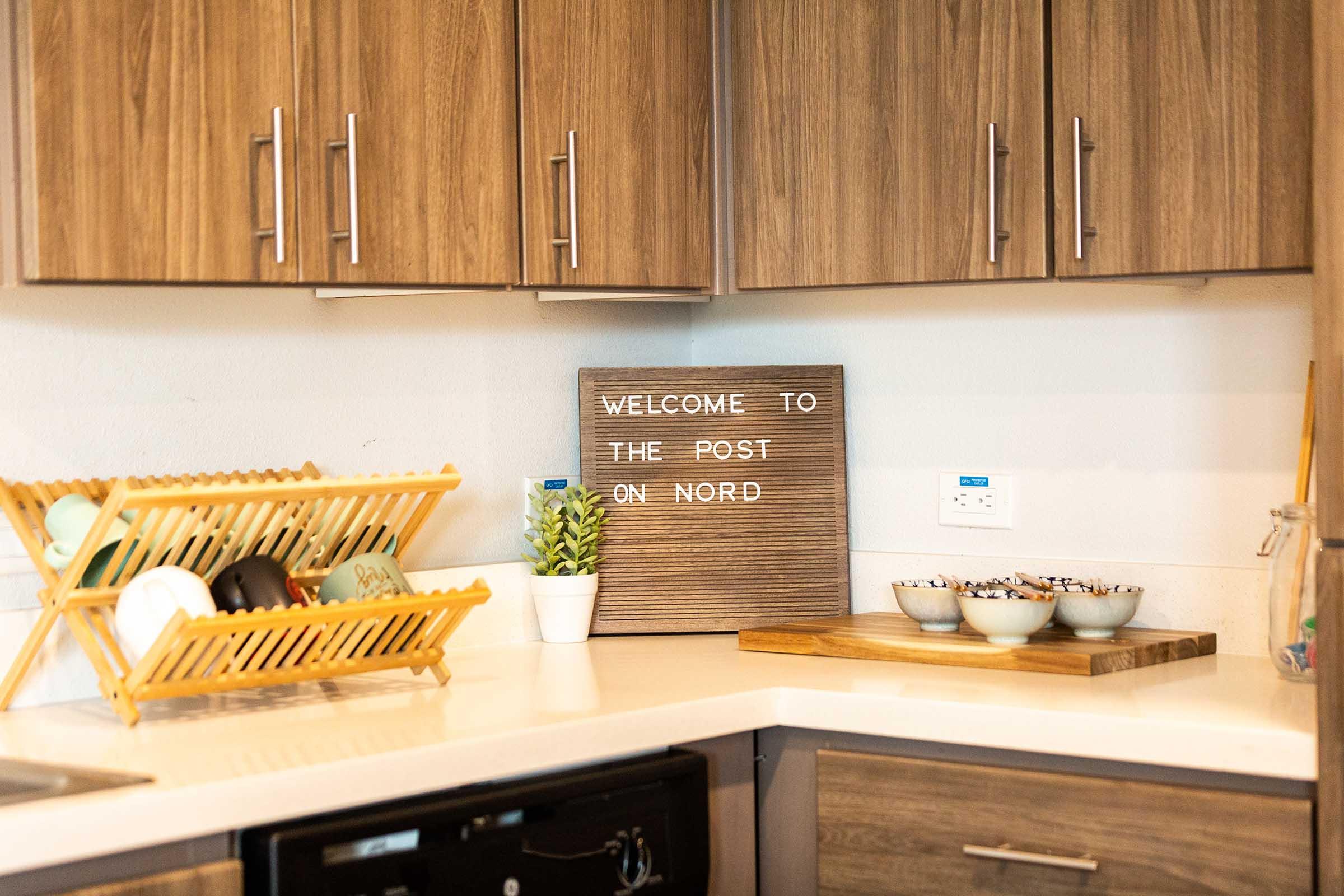 A modern kitchen with wooden cabinets and a countertop. A decorative sign reads "WELCOME TO THE POST ON NORD." There is a drying rack with dishware, a small plant, and bowls with snacks on a cutting board. An electric outlet is visible on the wall.