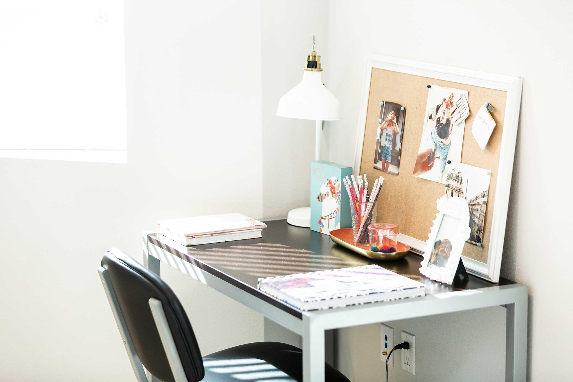 A modern desk with a black tabletop and a metal frame, featuring a lamp, decorative corkboard with photos, stationery, and a small picture frame. Nearby are colorful pens and a notepad. Natural light streams in through a window, casting soft shadows on the surface.