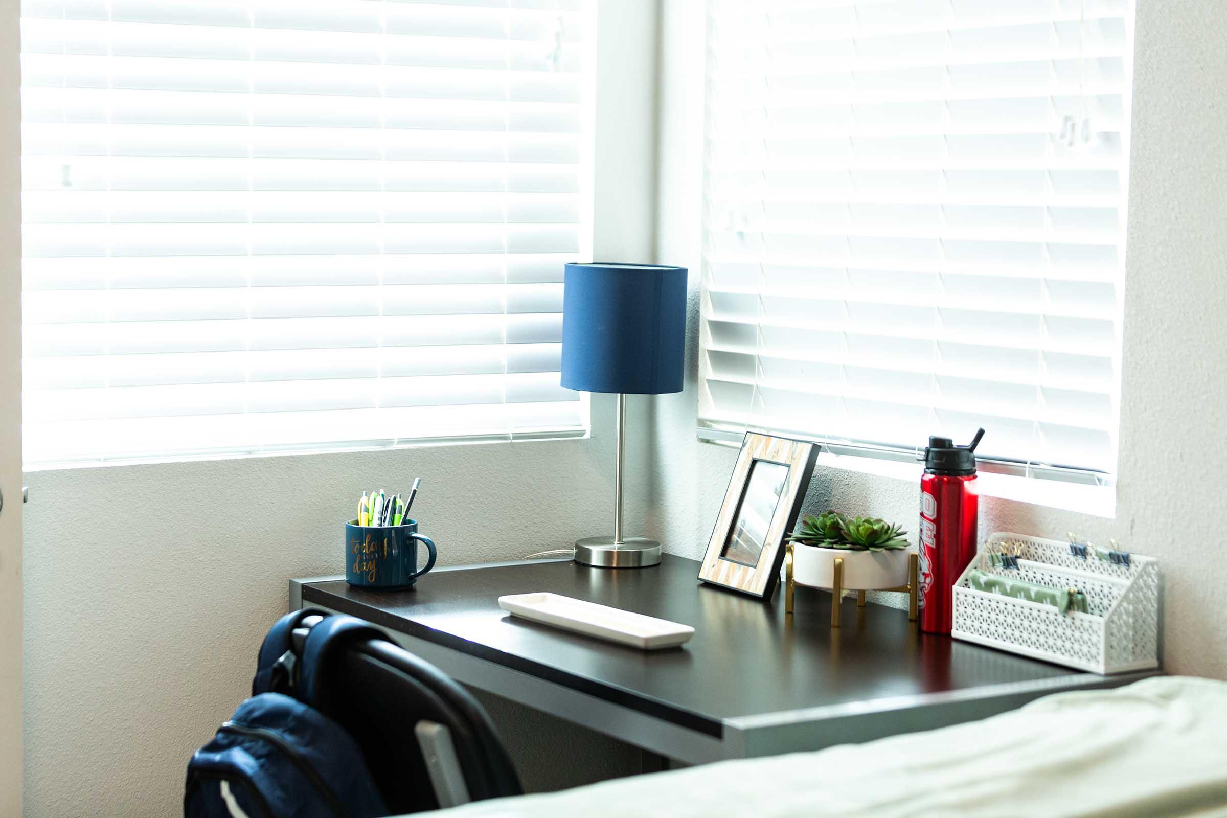 A corner desk by a window with blinds partially open. The desk features a blue lamp, a small framed photo, a plant in a pot, pens in a cup, a water bottle, and a tray with miscellaneous items. A chair is positioned beside the desk, and natural light brightens the space.