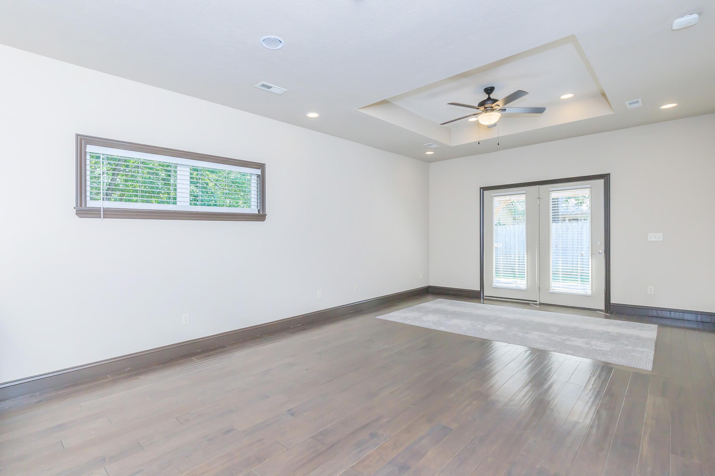 A spacious, empty living room featuring hardwood floors, a ceiling fan, and neutral wall colors. Large windows allow natural light, and a set of double doors leads to an outdoor area. A small area rug is placed on the floor, enhancing the room's welcoming atmosphere.