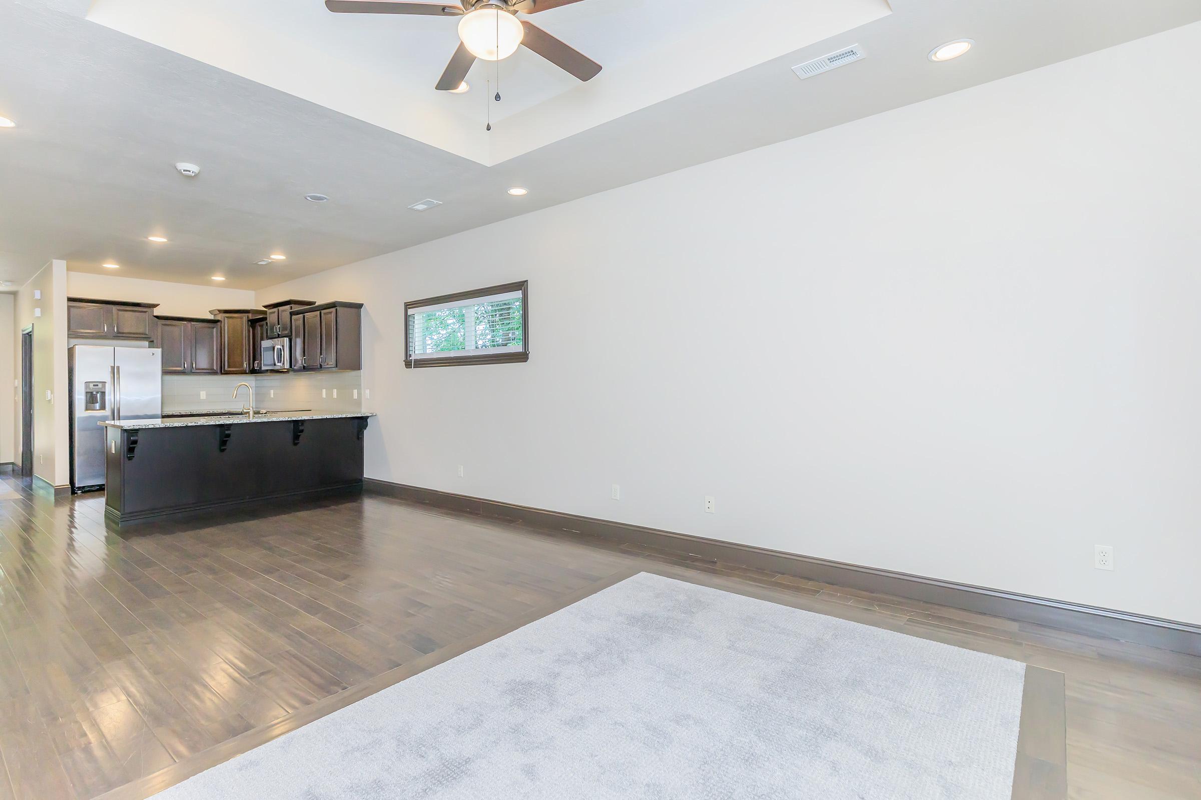 A spacious, empty living room featuring dark wood flooring, a ceiling fan, and light-colored walls. There's a large window on one side and an adjacent kitchen area with dark cabinetry and stainless steel appliances, creating an open and airy feel. A light-colored area rug is partially visible on the floor.