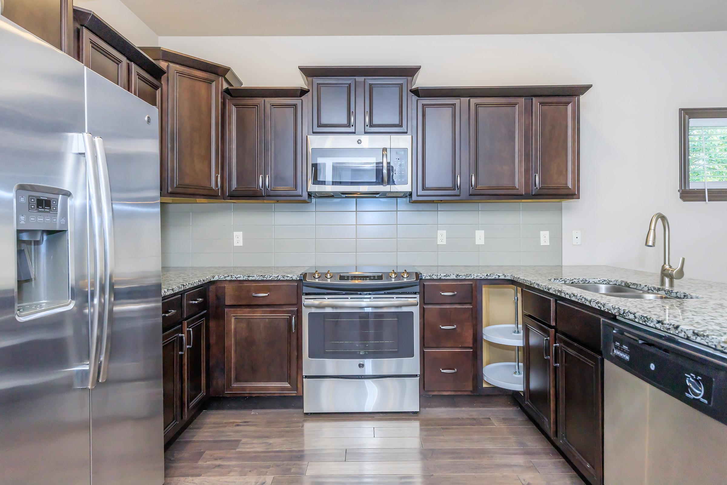 Modern kitchen with dark wooden cabinetry, a stainless steel refrigerator, and a microwave above a stovetop. The countertop is made of gray granite, and there are details like a circular storage unit. The flooring is hardwood, and the walls are painted in a light color. Natural light comes from a nearby window.