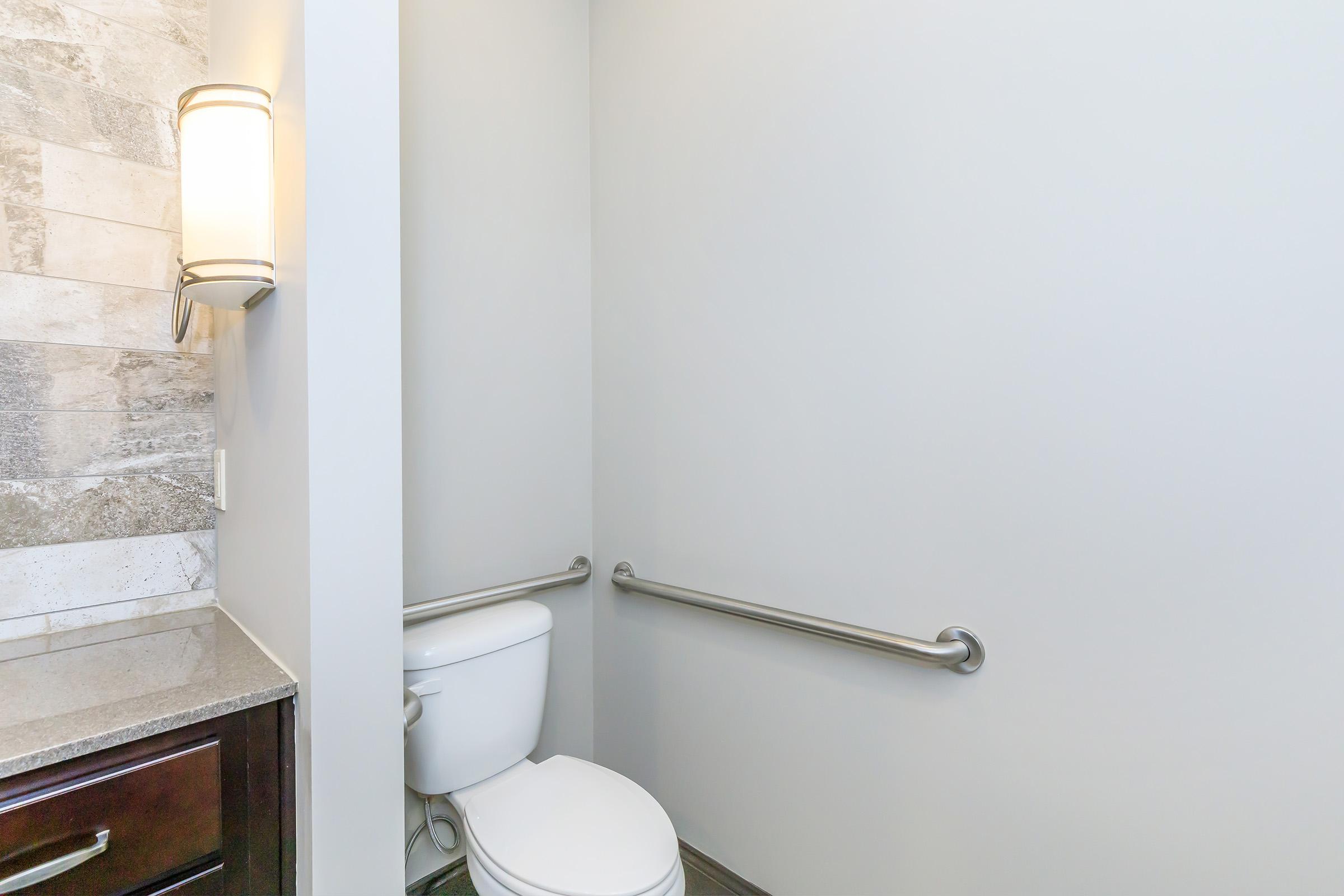 A clean bathroom stall featuring a white toilet, grab bars on the walls for accessibility, and a light-colored wall with textured tiles. The flooring is dark, and there is a countertop with a sink area visible in the corner.