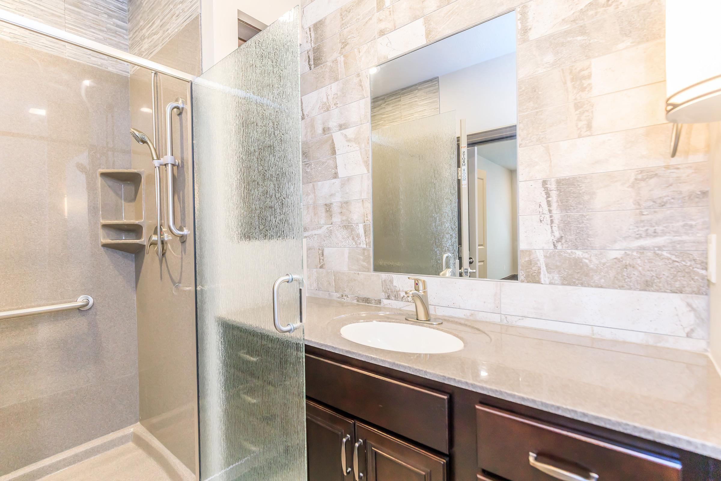Modern bathroom featuring a glass shower enclosure with a built-in bench, contemporary gray tiles, and a sleek granite vanity. A large mirror above the sink reflects the stylish design, and there are brushed metal fixtures complementing the overall aesthetic of the space.