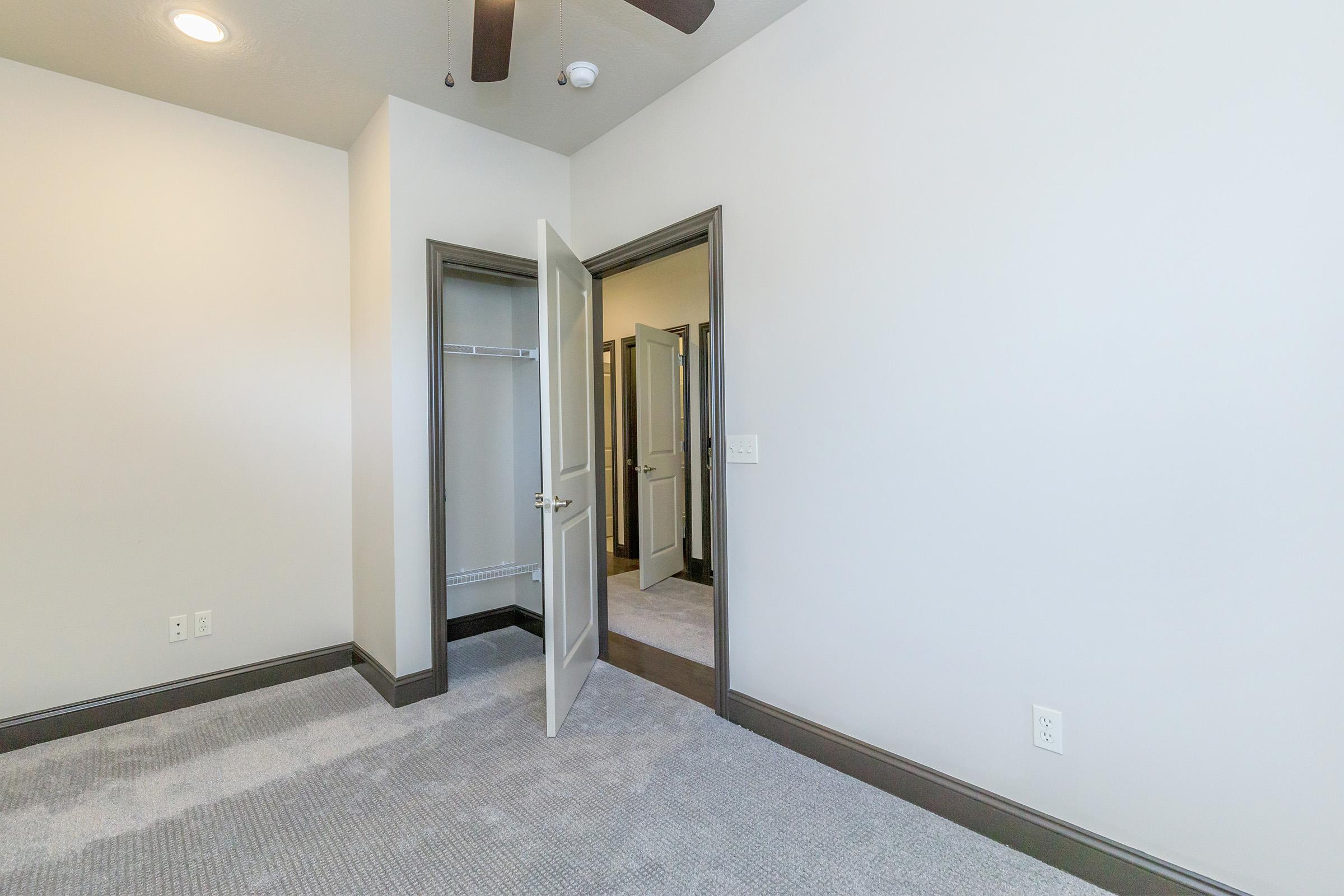 A small room with light gray walls and carpet. There are two open doors leading to closets, and a ceiling fan is visible. The overall lighting is soft and inviting, creating a spacious feel in the interior.