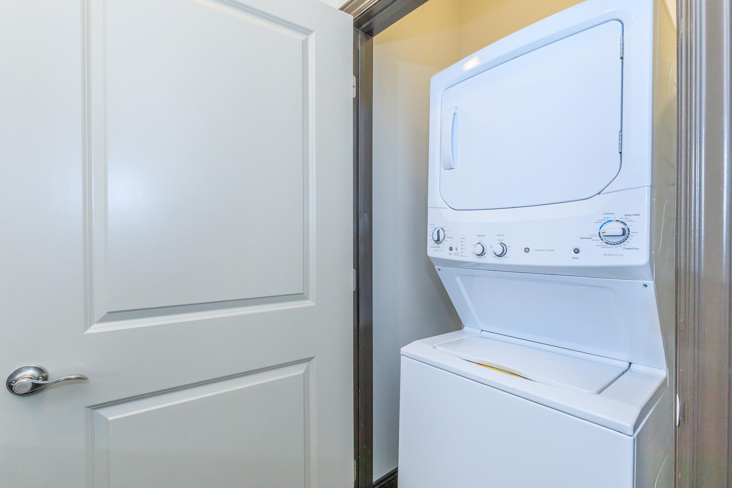 A white stacked washer and dryer unit is positioned in a narrow laundry space beside a closed door. The appliance features dials and settings for washing and drying, and the space is well-lit with a neutral-colored wall.