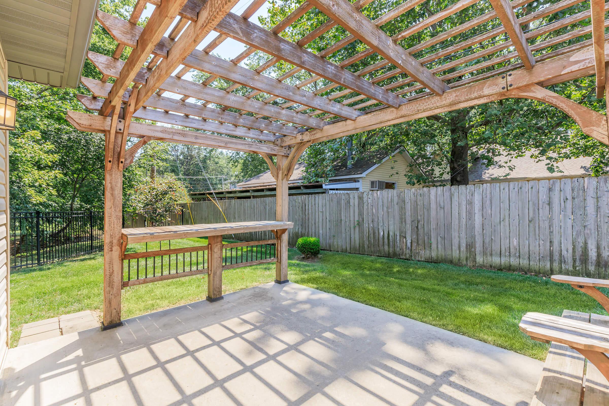 A wooden pergola with a slatted roof, providing partial shade, stands in a backyard with a lawn. There is a small green bush and a concrete patio beneath the pergola. In the background, a wooden fence encloses the yard, and trees are visible, creating a peaceful outdoor setting.