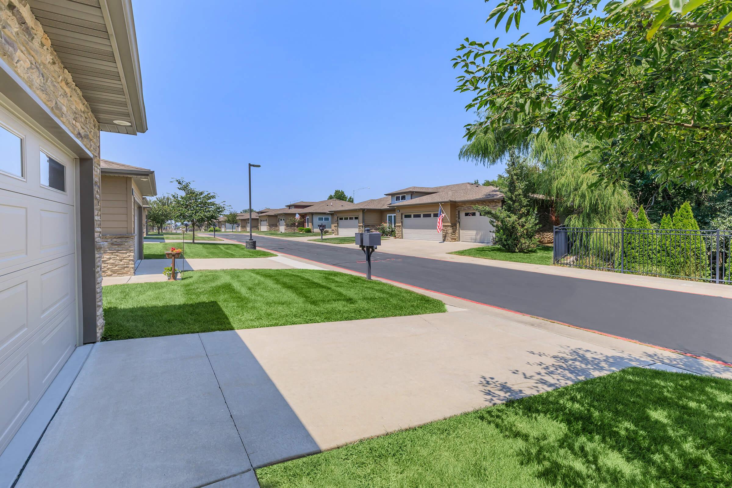 A serene residential street lined with well-maintained homes. Neatly trimmed green grass and concrete walkways frame the properties. A mailbox stands at the edge of a driveway, and trees provide shade. The sky is clear and blue, creating a pleasant atmosphere in this suburban neighborhood.