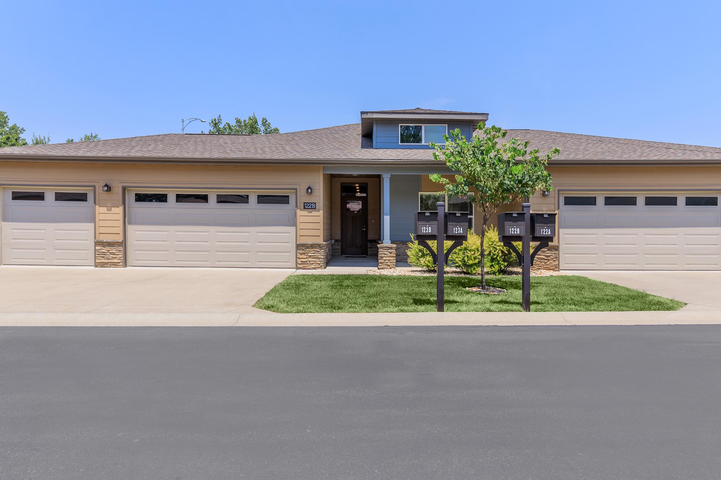 Residential building with a modern design featuring a wide driveway, three garage doors, and a front entrance. There are mailbox stands with multiple mailboxes and well-maintained landscaping including a small tree and grassy areas. The sky is clear and blue.