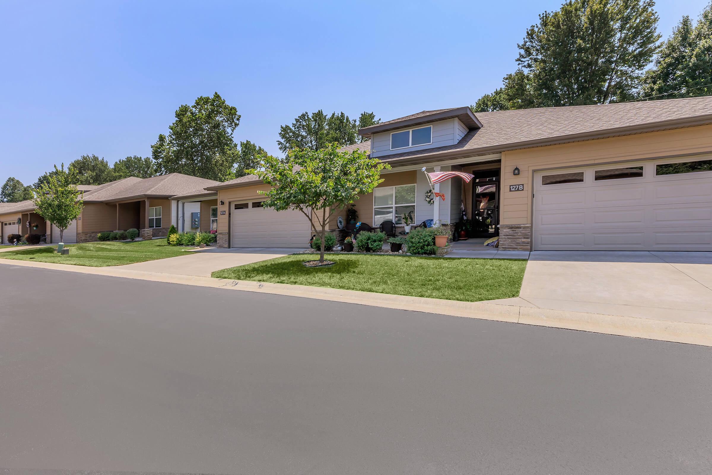 A view of a suburban neighborhood showing several one-story houses with well-maintained lawns. Trees and shrubs are present, along with driveways leading to garages. The sky is clear and blue, suggesting a sunny day, and there are decorative elements like plants on the porches.
