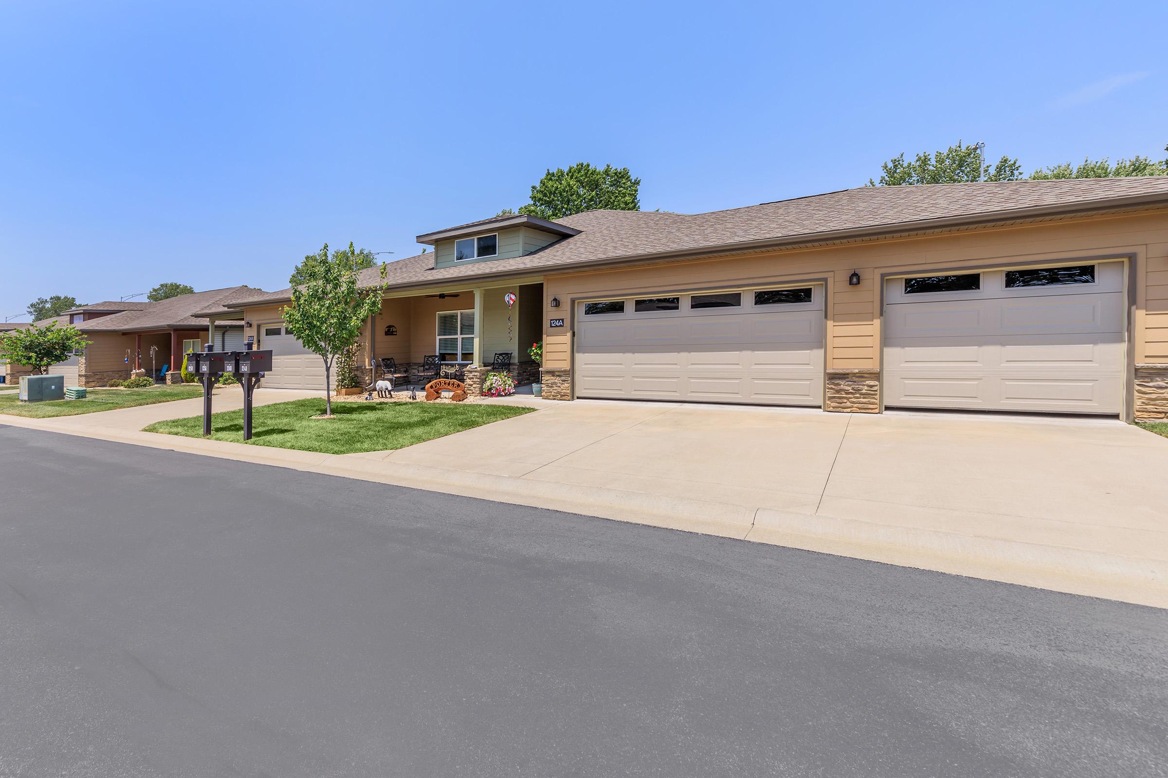 A suburban street view showing a row of single-story homes. Each home features a garage with a paved driveway, neatly trimmed lawns, and decorative plants. The sky is clear and blue, adding to the pleasant atmosphere of the neighborhood.