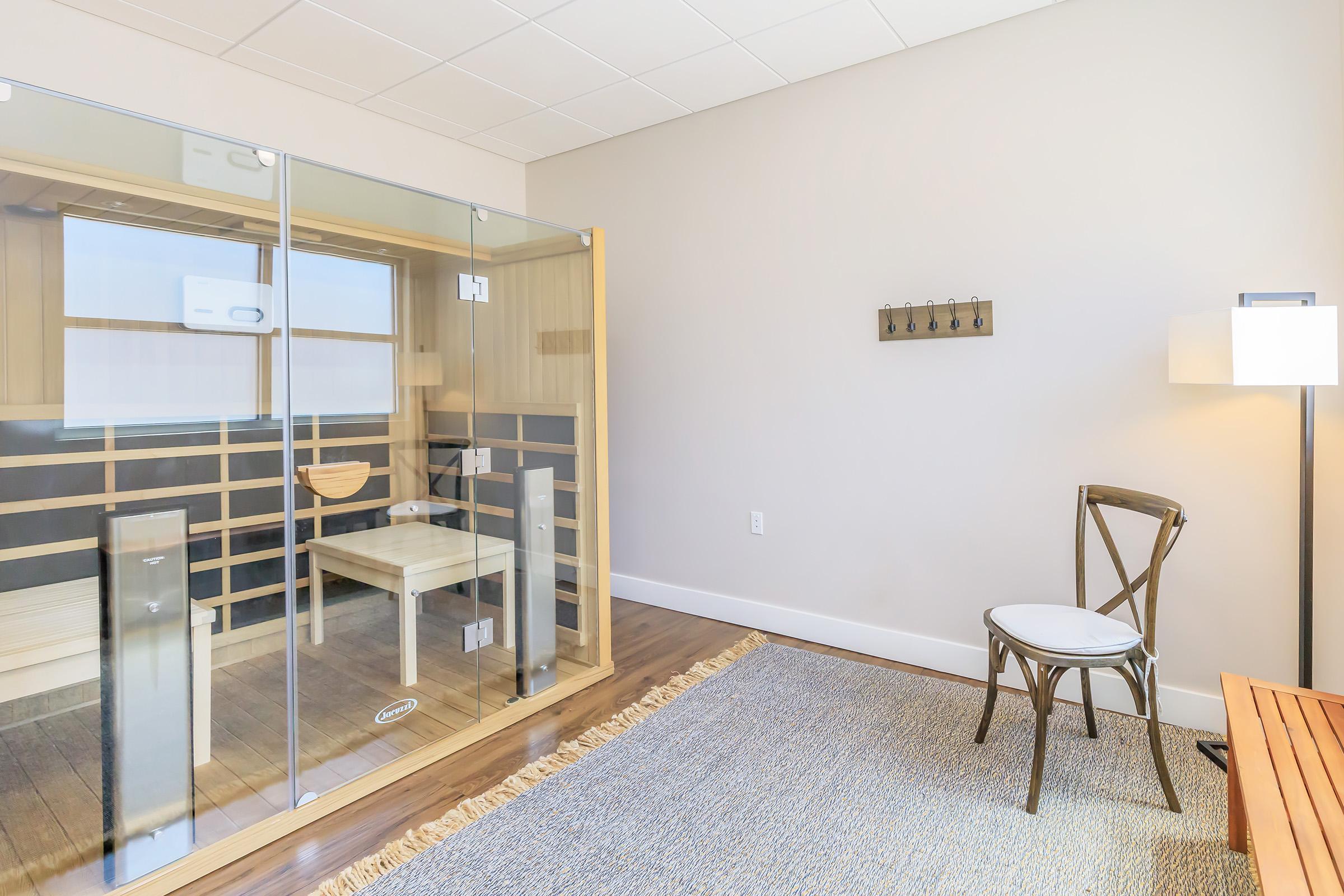 A serene indoor space featuring a glass-enclosed sauna on one side, a small wooden table inside the sauna, and a simple chair placed on a textured rug. A lamp stands beside the chair, and a wall-mounted coat rack is visible. The neutral-colored walls and warm wood tones create a calming atmosphere.