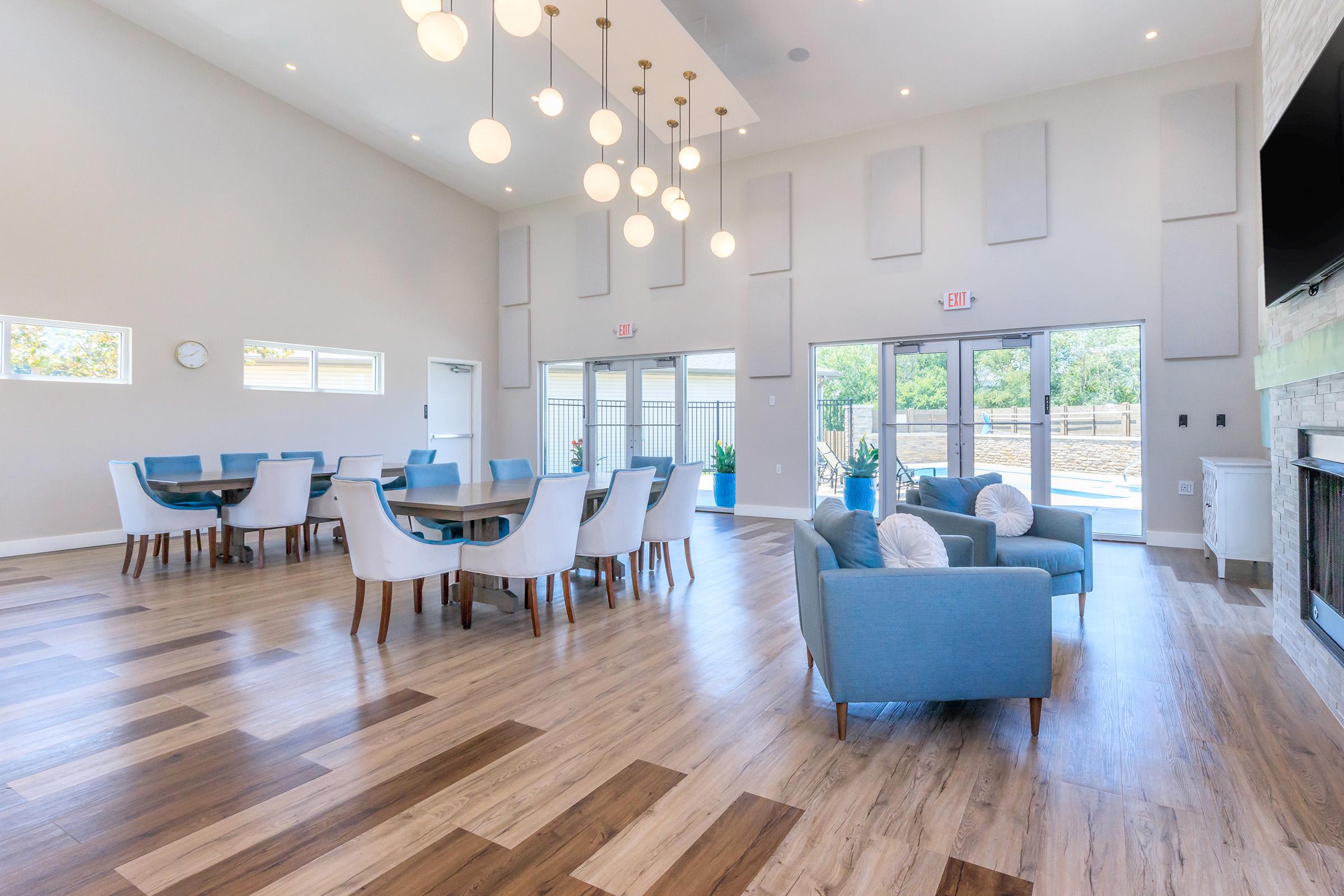 Interior view of a spacious community room featuring a large, round dining table surrounded by chairs, and several seating areas with blue armchairs. The room has large windows allowing natural light, a modern light fixture, and wood flooring that adds warmth to the space.