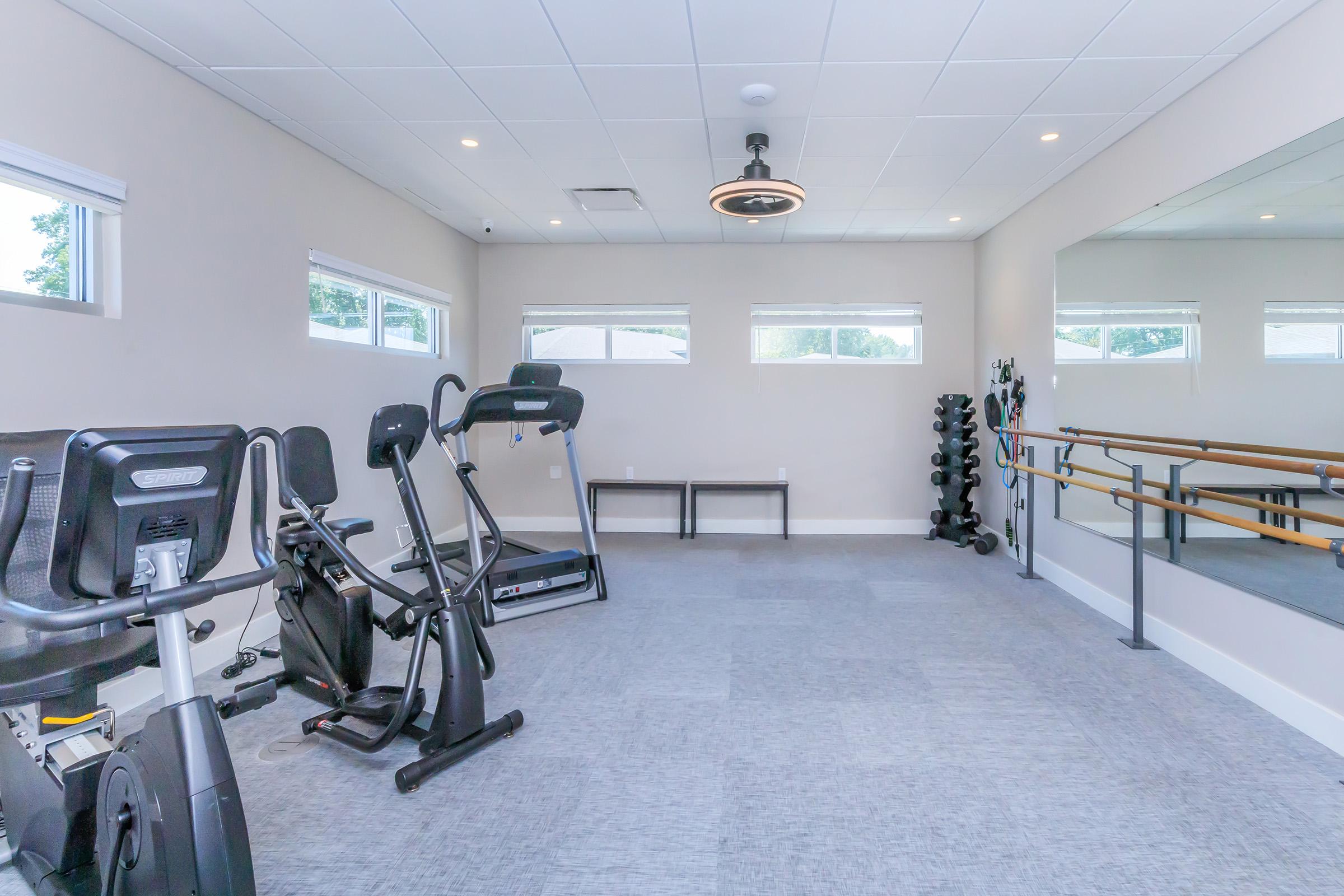 A spacious exercise room featuring two fitness machines, a mirrored wall, and a barre. Natural light streams in through the windows, illuminating the gray carpet and minimalistic decor. Dumbbells are neatly arranged in a corner, creating an inviting atmosphere for workouts.